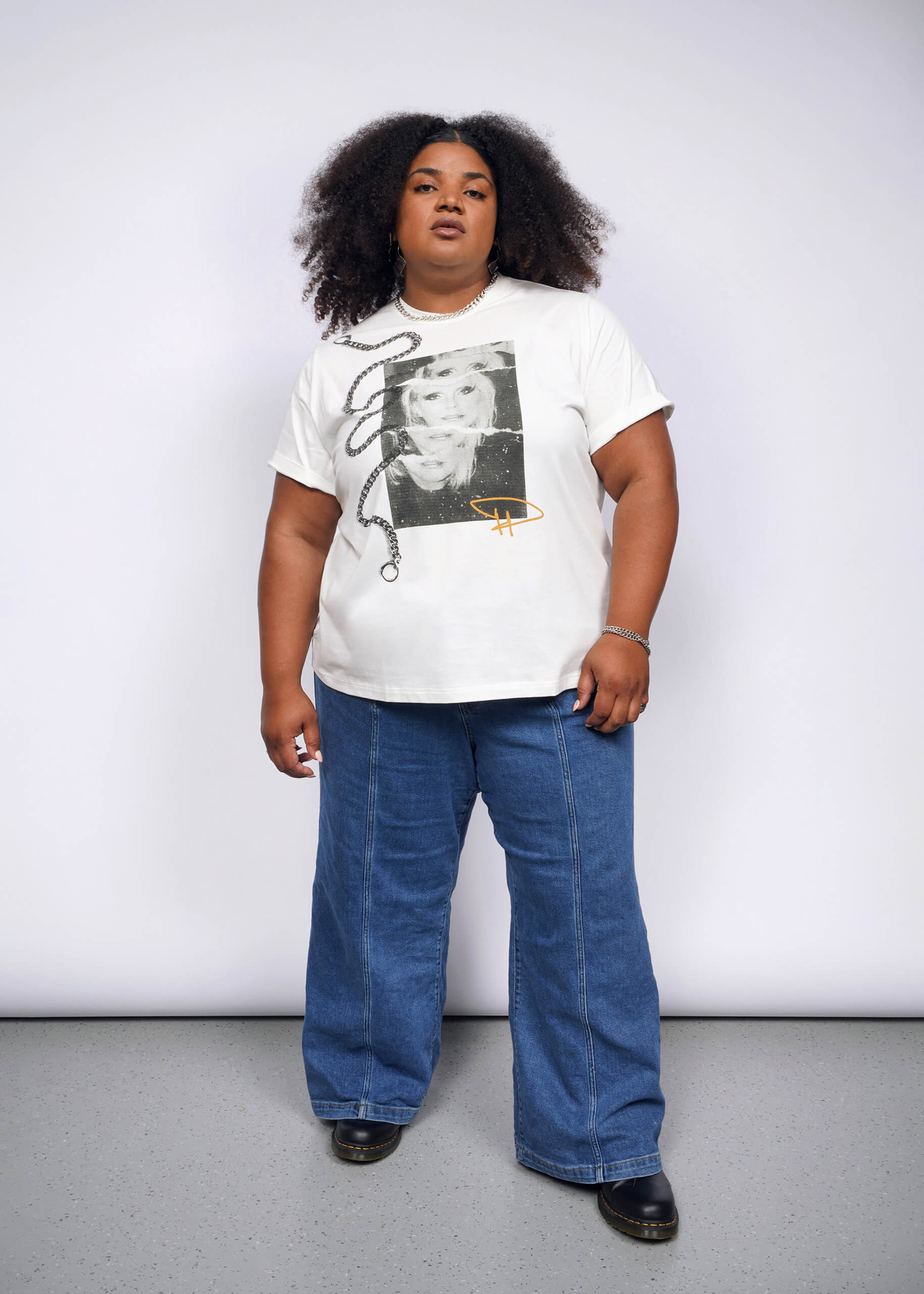 1X= A curly-haired person, embodying a punk aesthetic, stands confidently against a plain background. Theyre wearing the Debbie Harry X Wildfang Graphic Tee with blue jeans and black shoes, exuding the charisma of Debbie Harry.