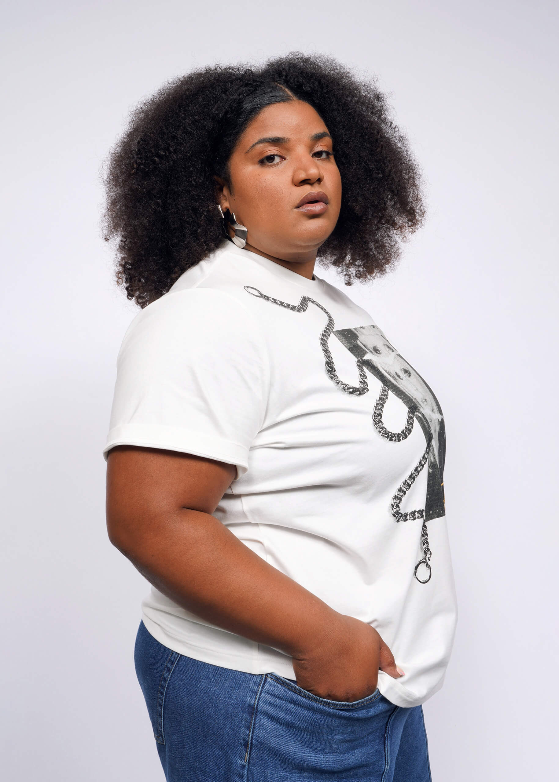 1X= A person with curly hair channels a Debbie Harry vibe, wearing the Debbie Harry X Wildfang Graphic Tee and blue jeans. They stand confidently against a plain background, looking directly at the camera with one hand in their pocket, exuding a subtle punk aesthetic.