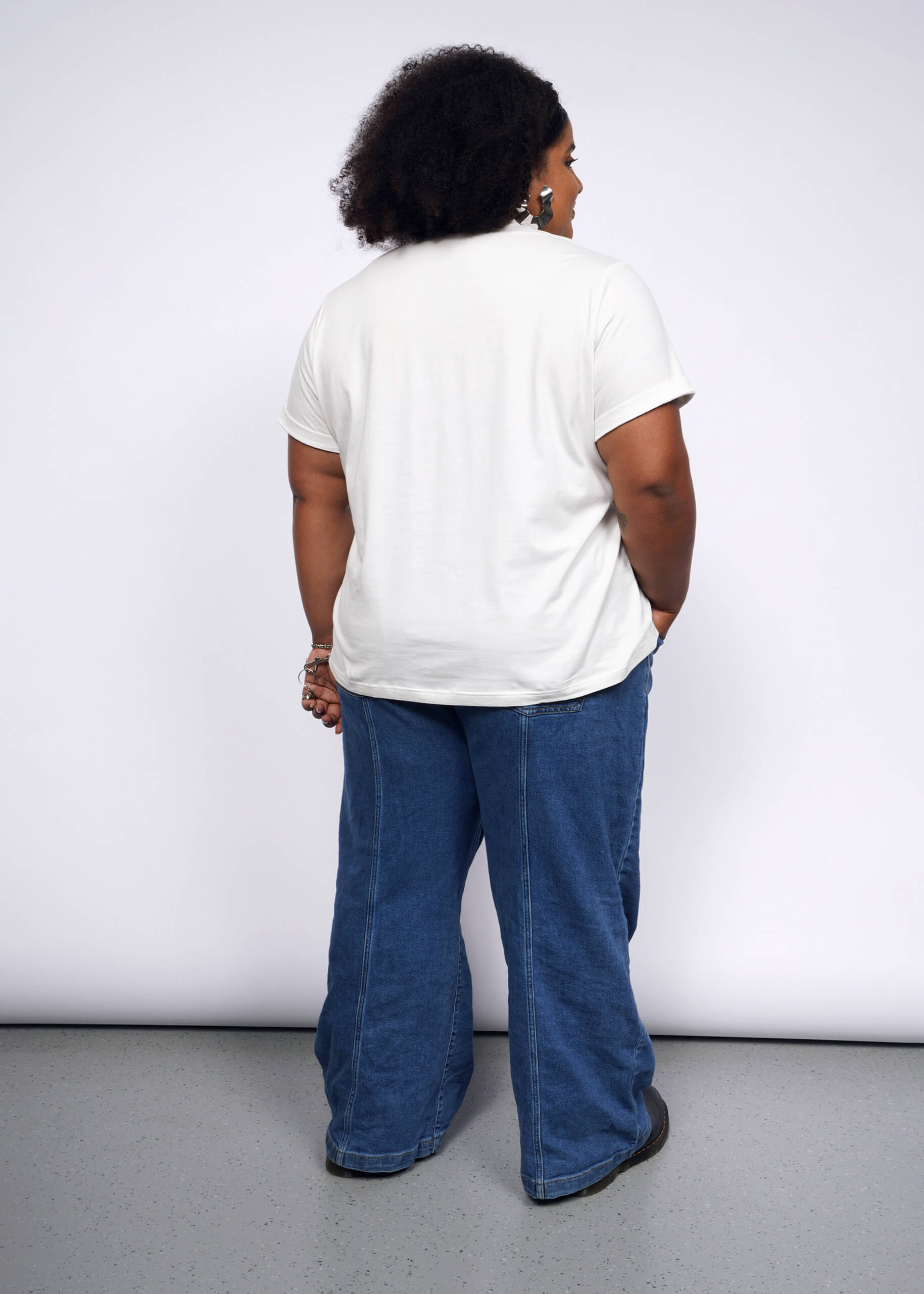 1X=A person with curly hair faces away from the camera against a white backdrop, embracing a punk vibe. Theyre wearing the Debbie Harry X Wildfang Graphic Tee and wide-legged blue jeans, their hands relaxed by their sides.