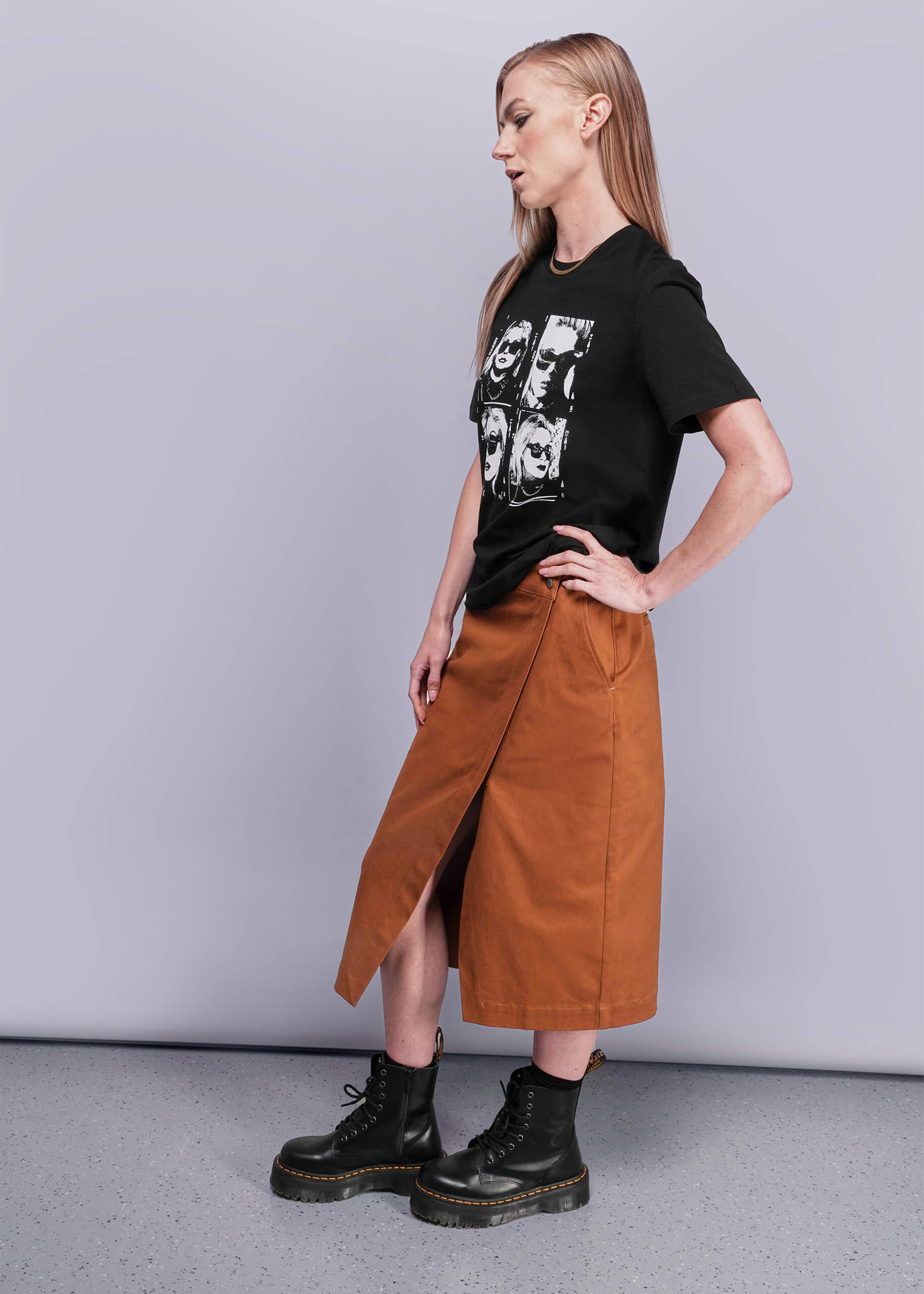 S=Person wearing a black t-shirt with graphic design, brown skirt, and black boots on a plain background