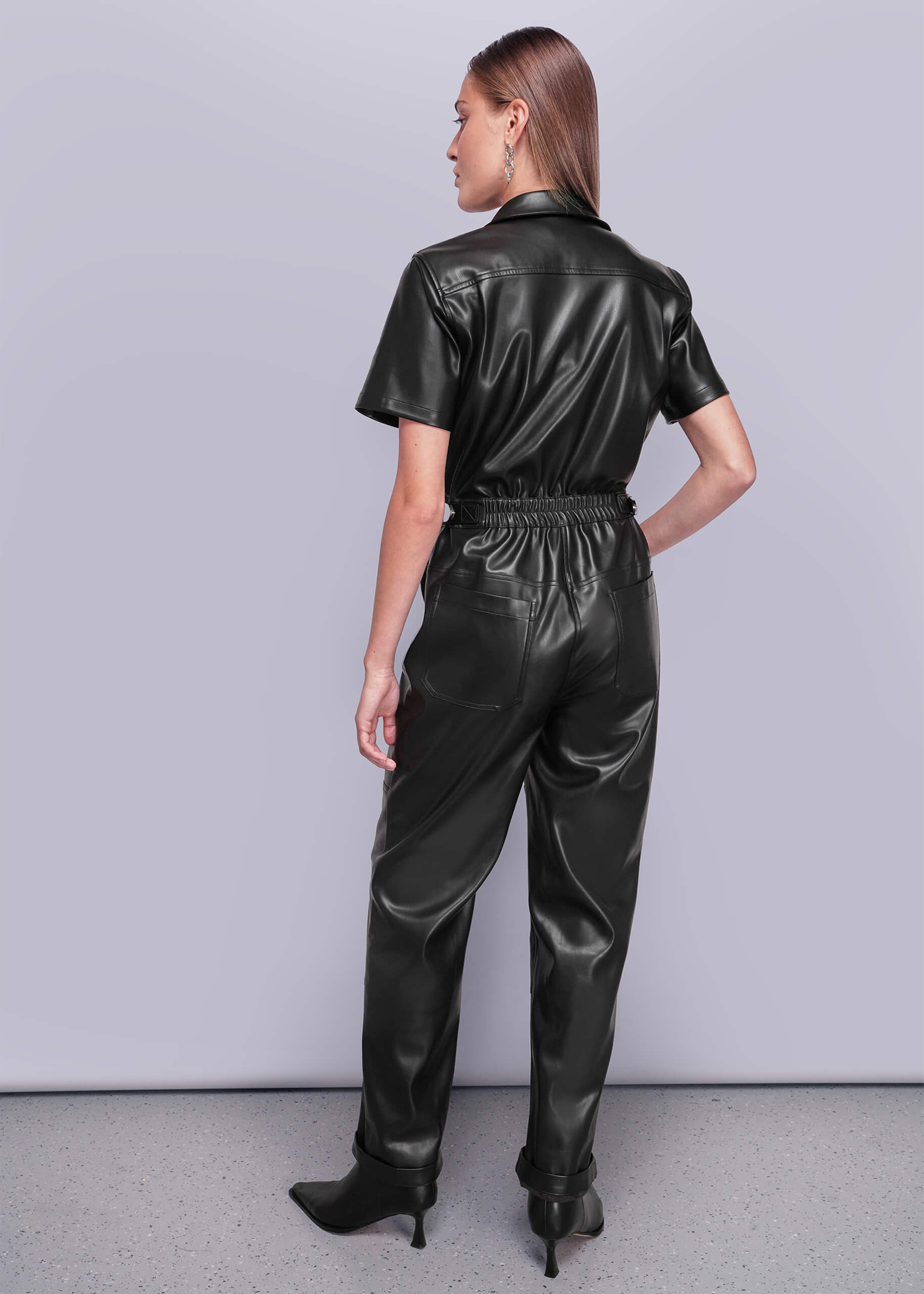 S=Back view of person wearing a black vegan leather jumpsuit against a plain background
