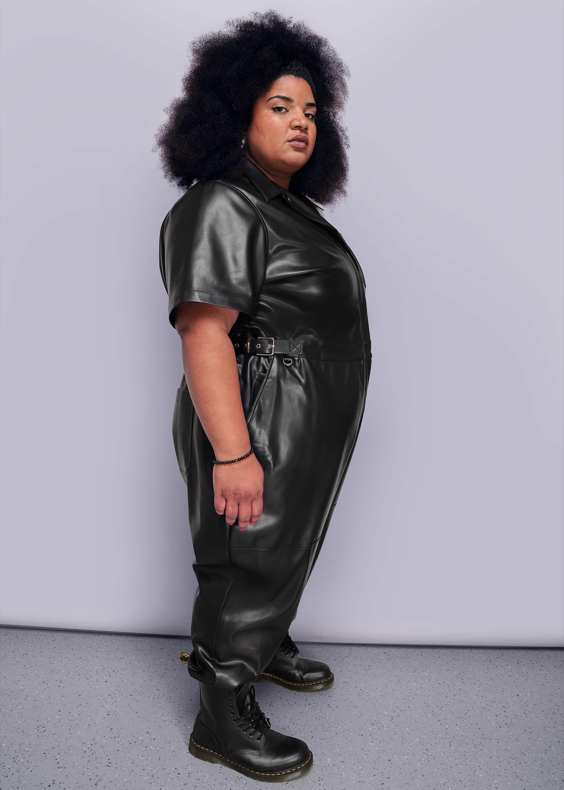 1X=Side view of person wearing a black vegan leather jumpsuit against a plain background