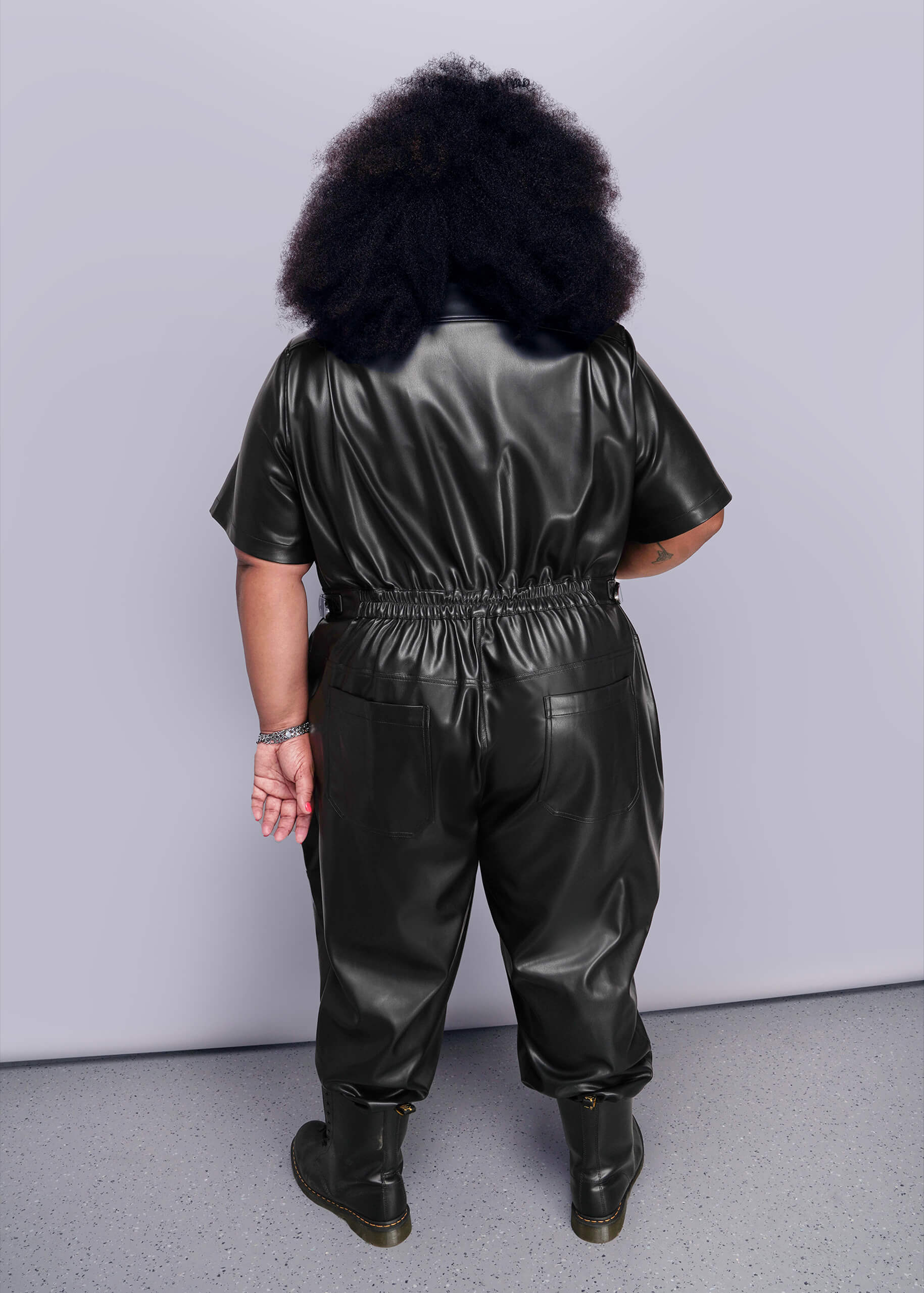 1X=Person wearing a black vegan leather jumpsuit against a plain background