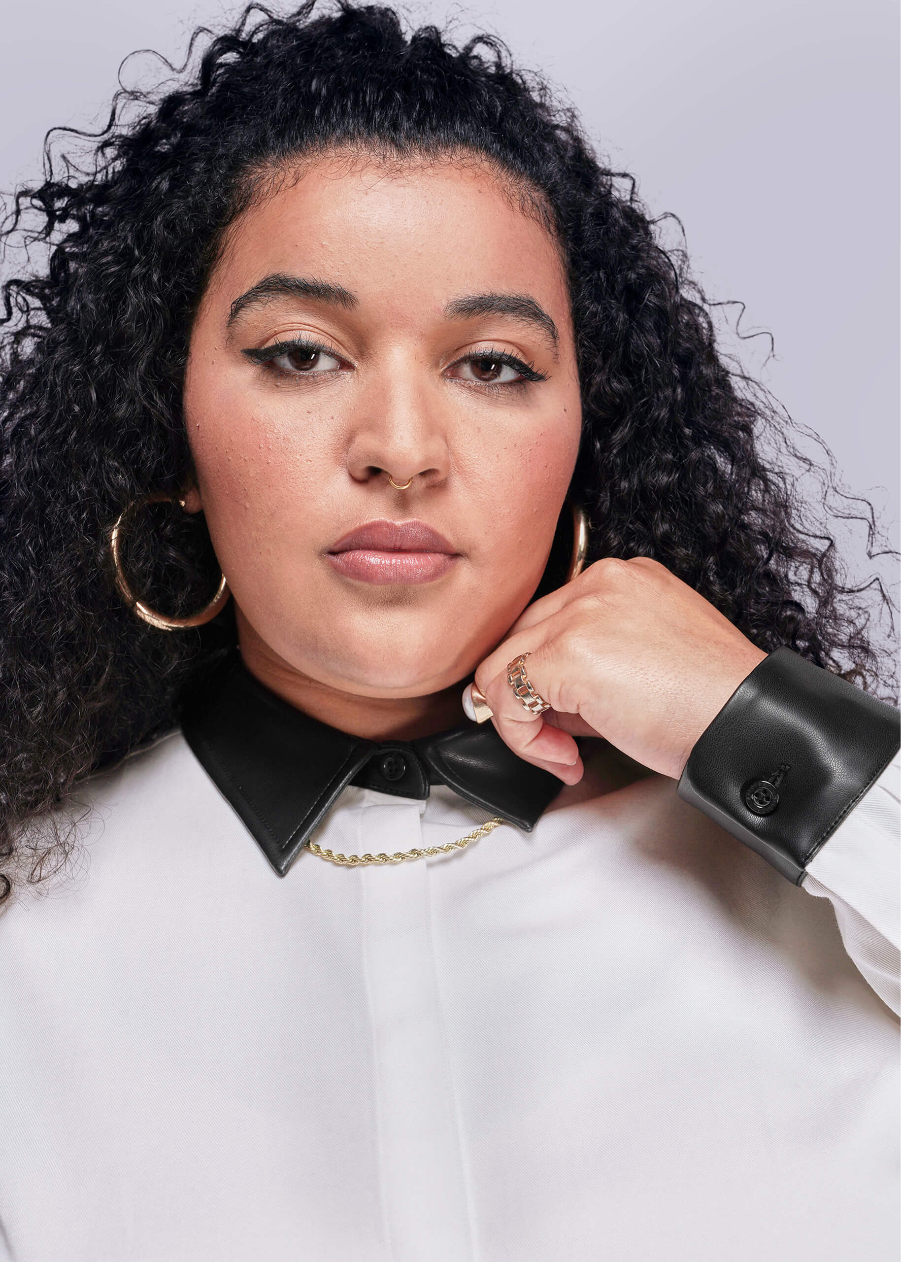 1X=Woman wearing a white button up with a black vegan leather collar, and gold hoop earrings on a light gray background