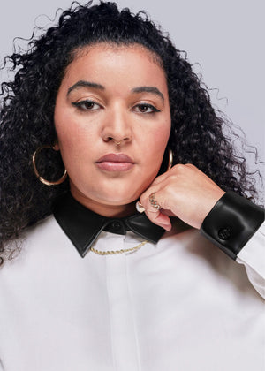 1X=Woman wearing a white button up with a black vegan leather collar, and gold hoop earrings on a light gray background