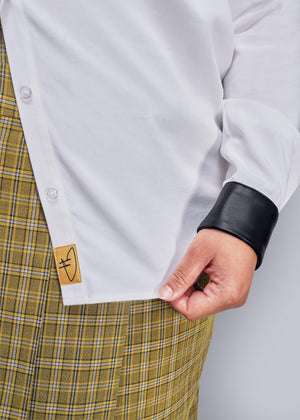 1X=Close-up of a person wearing a white shirt with a black vegan leather cuff and yellow plaid skirt.