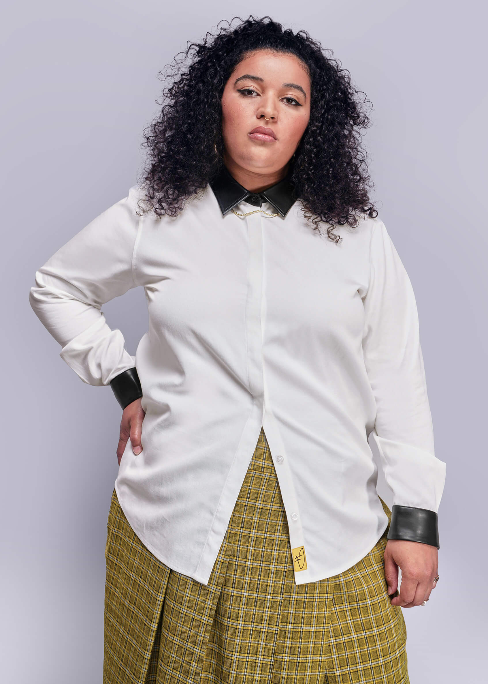 1X=Person wearing a white shirt with black vegan leather cuffs and a yellow plaid skirt on a light gray background