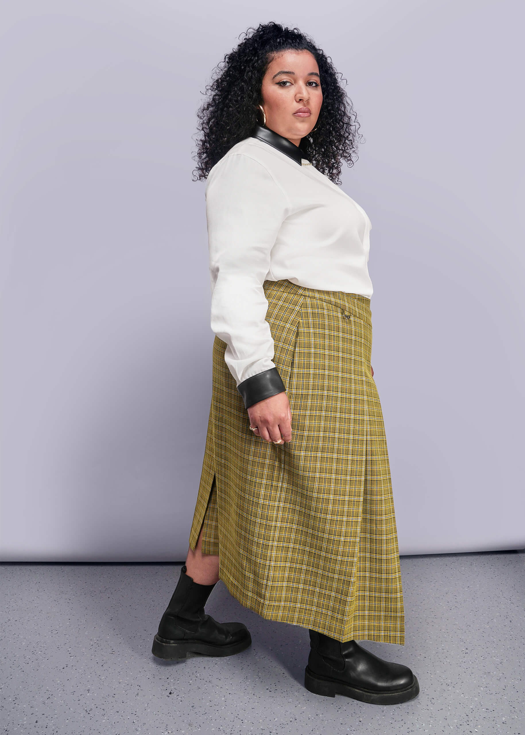 1X=Side view of person wearing a white button up shirt, yellow plaid skirt, and black boots against a plain background