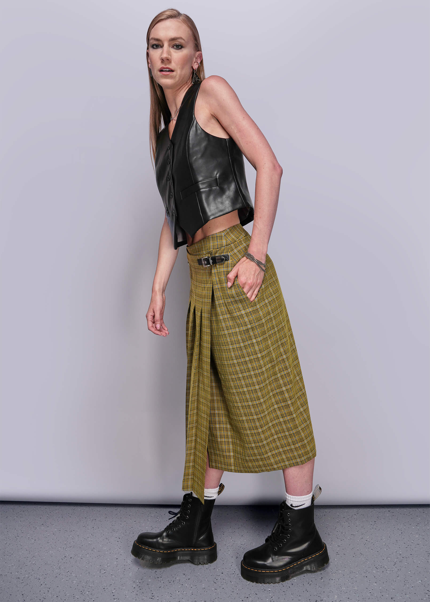 S=Side view of person wearing a black leather vest, yellow plaid skirt, and black boots against a plain background