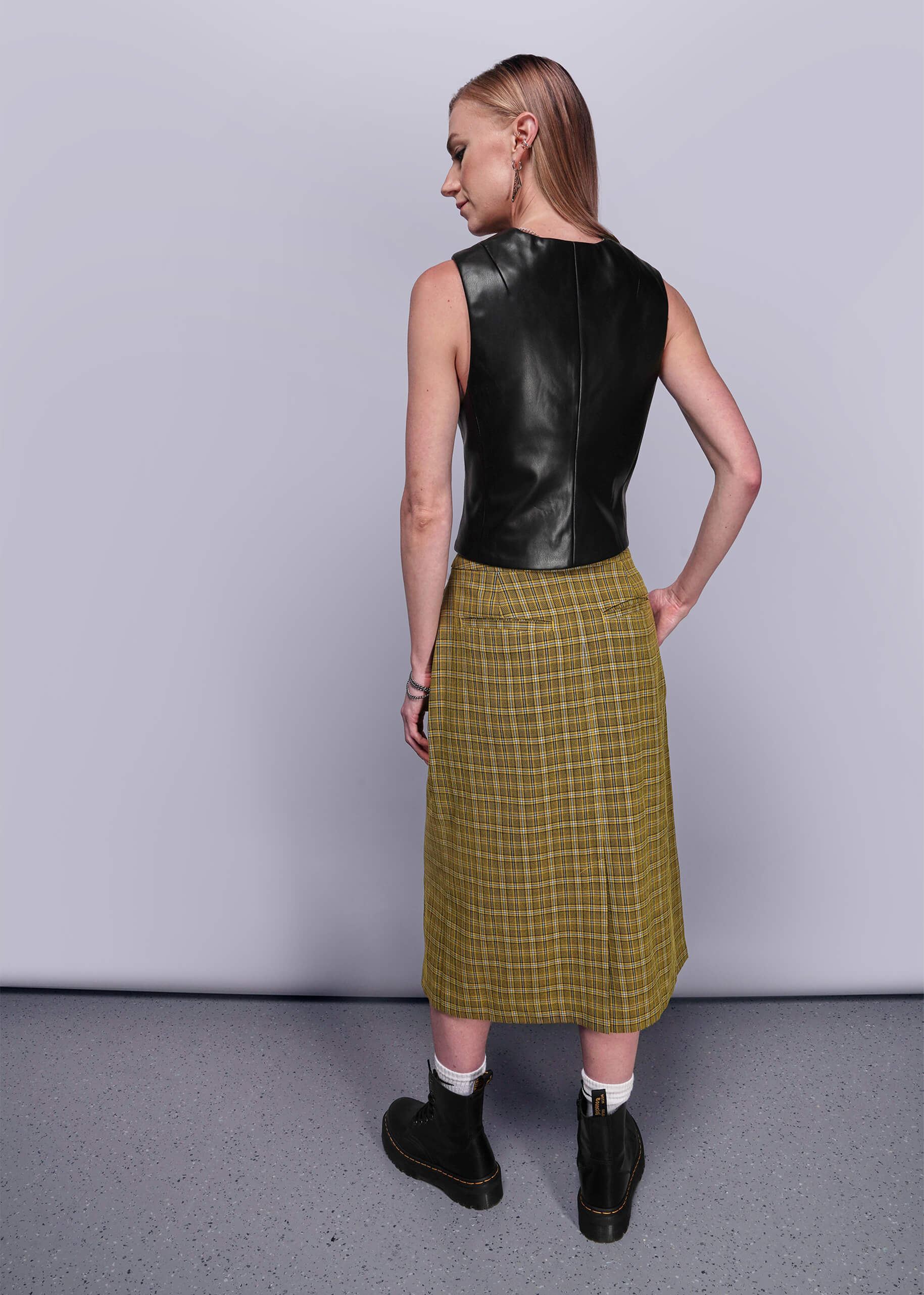 S=Back view of person wearing a black leather vest and yellow plaid skirt against a plain background