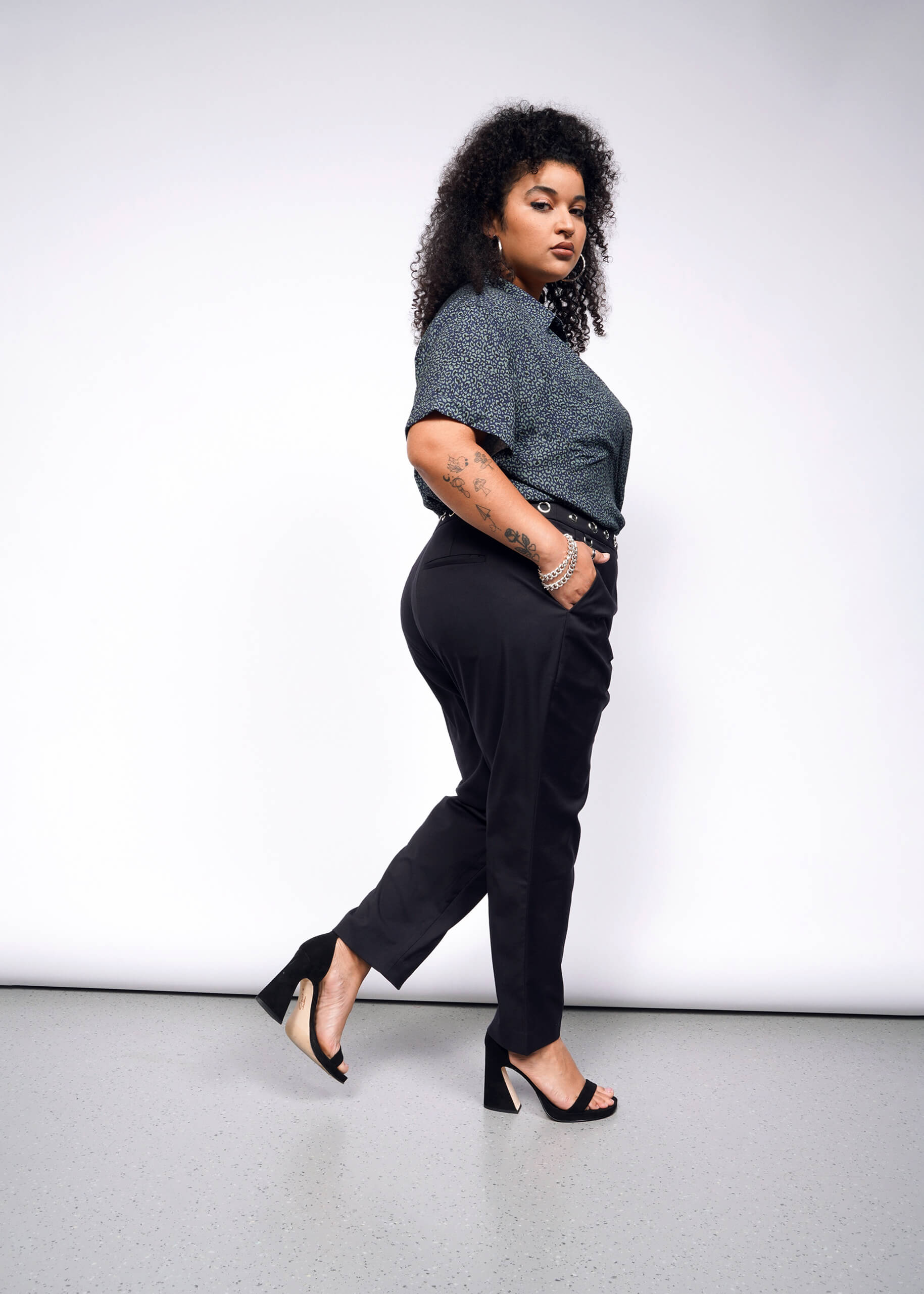 1X=With curly hair, a person channels a punk vibe, confidently posing in a dark speckled top, Debbie Harry X Wildfang Slim Crop Pant, and high heels. The plain white background highlights their bold look as they stride forward.