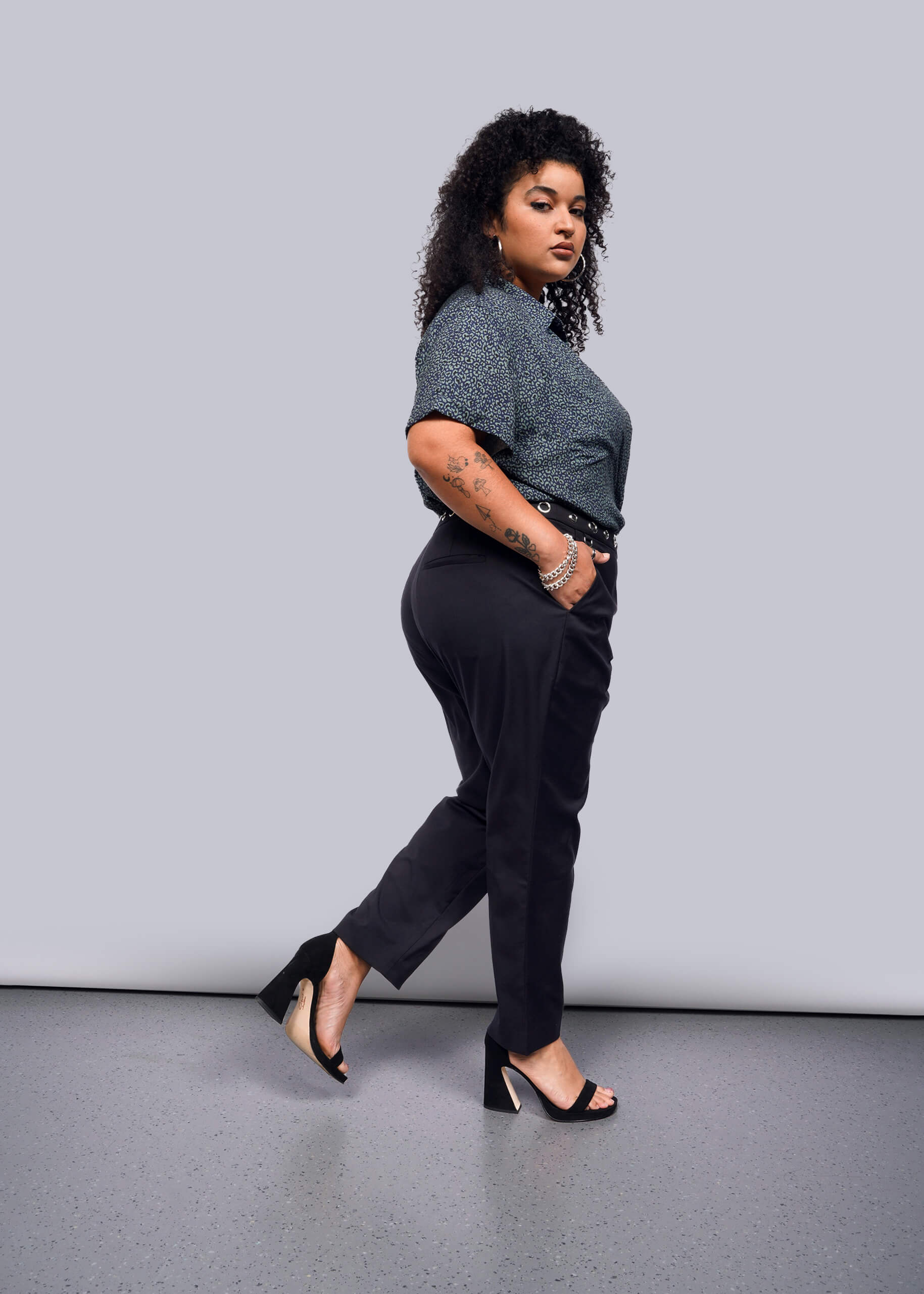 1X=With curly hair, a person channels a punk vibe, confidently posing in a dark speckled top, Debbie Harry X Wildfang Slim Crop Pant, and high heels. The plain white background highlights their bold look as they stride forward.