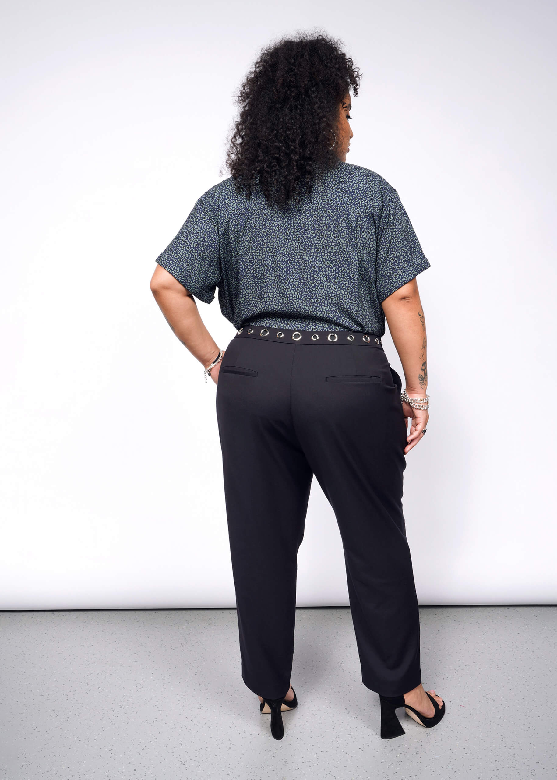 1X= With curly hair exuding a subtle punk vibe, they stand out in a blue-patterned Wildfang collection shirt and Debbie Harry X Wildfang Slim Crop Pant. Black heels and bracelets on their wrist add an edgy flair against the plain background.