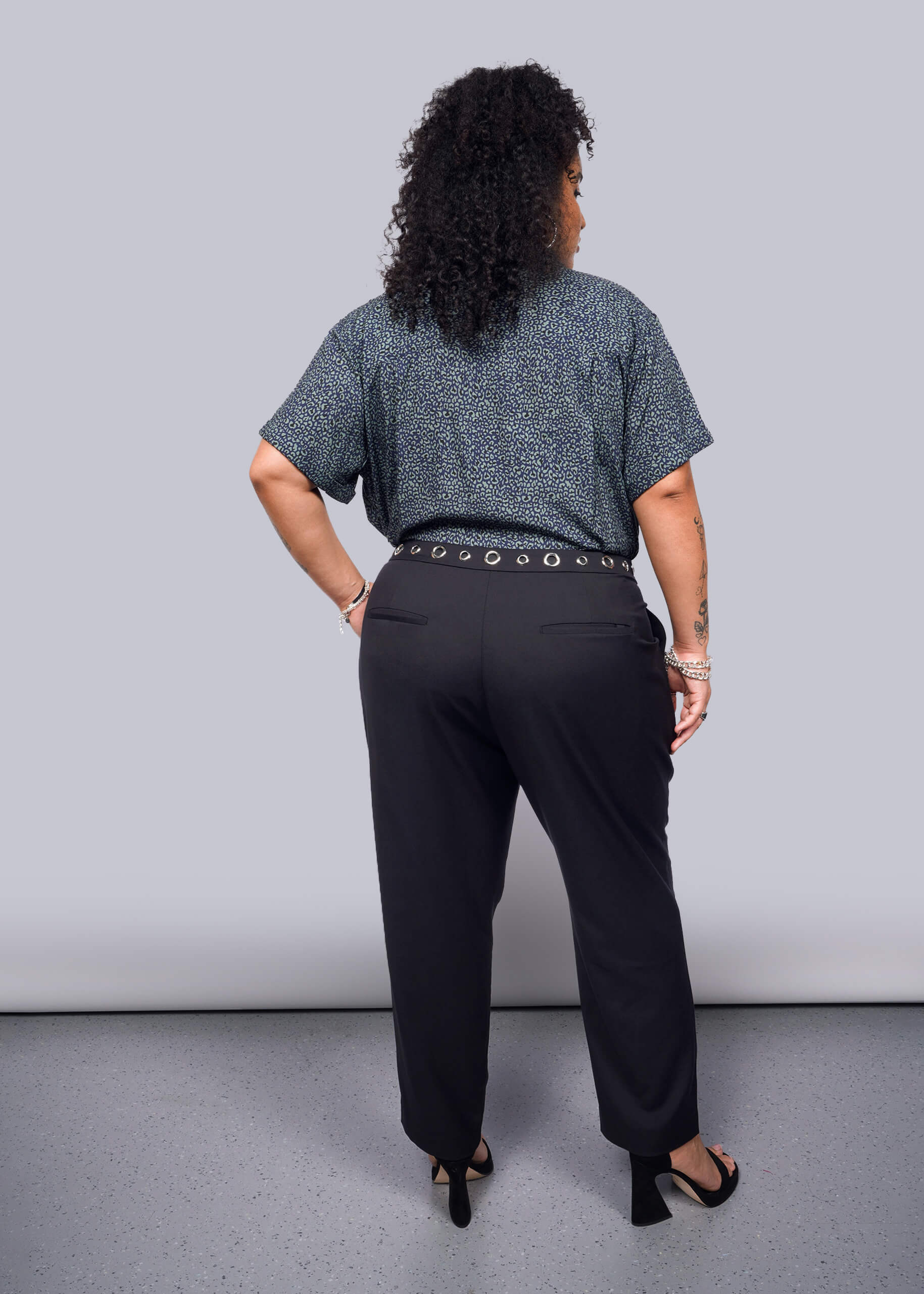 1X= With curly hair exuding a subtle punk vibe, they stand out in a blue-patterned Wildfang collection shirt and Debbie Harry X Wildfang Slim Crop Pant. Black heels and bracelets on their wrist add an edgy flair against the plain background.