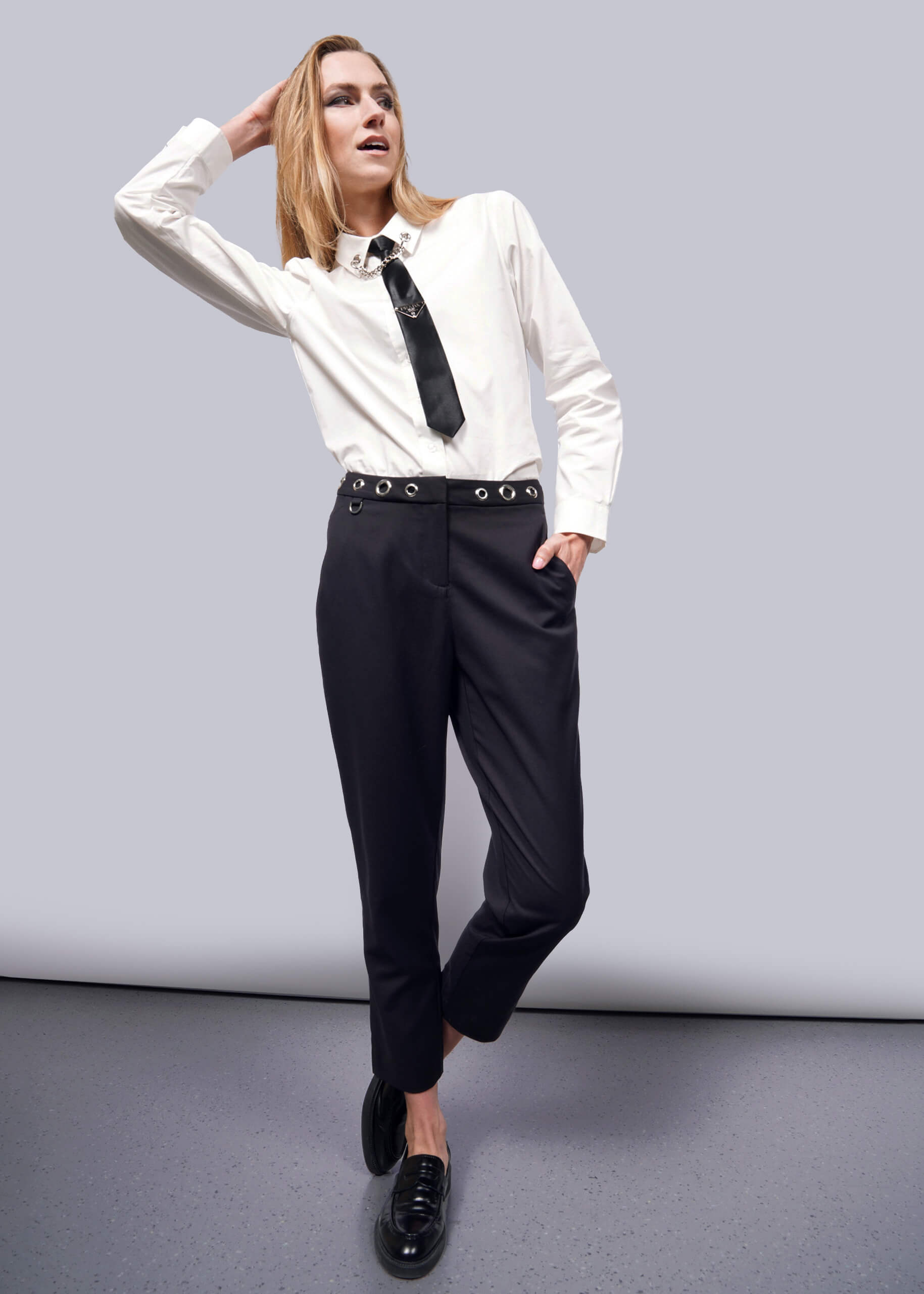S=Sporting the Debbie Harry X Wildfang Slim Crop Pant, a person channels punk style with a white shirt, black tie, and grommet-detailed black pants. They stand against a plain backdrop in black shoes, one hand on their head and the other in their pocket.