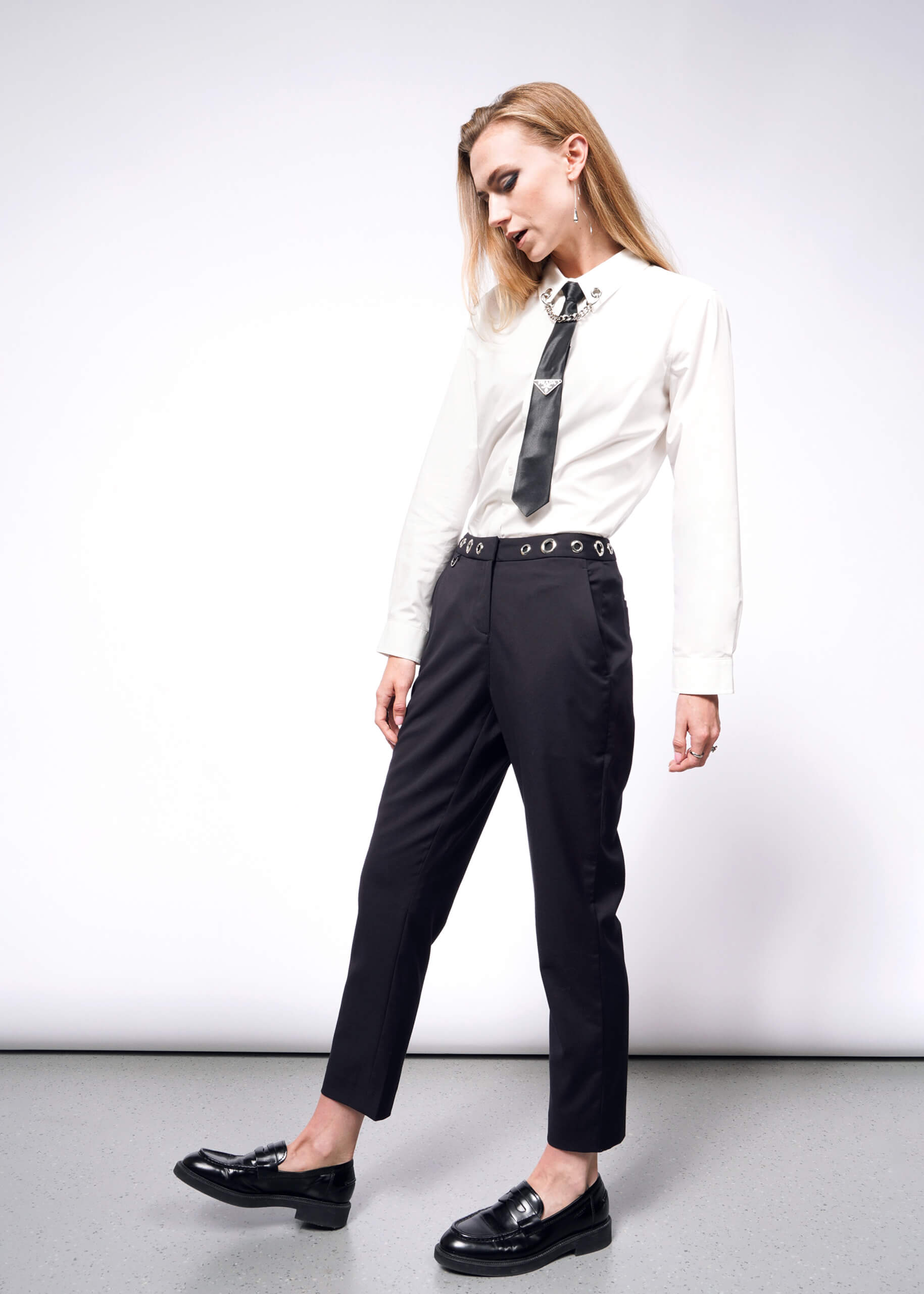 S= A person with long hair channels a punk aesthetic akin to Debbie Harry, standing relaxed against a plain backdrop. They wear a white shirt, black tie, and black loafers from the Wildfang collection, paired with the Debbie Harry X Wildfang Slim Crop Pant. Their head is slightly tilted downwards.