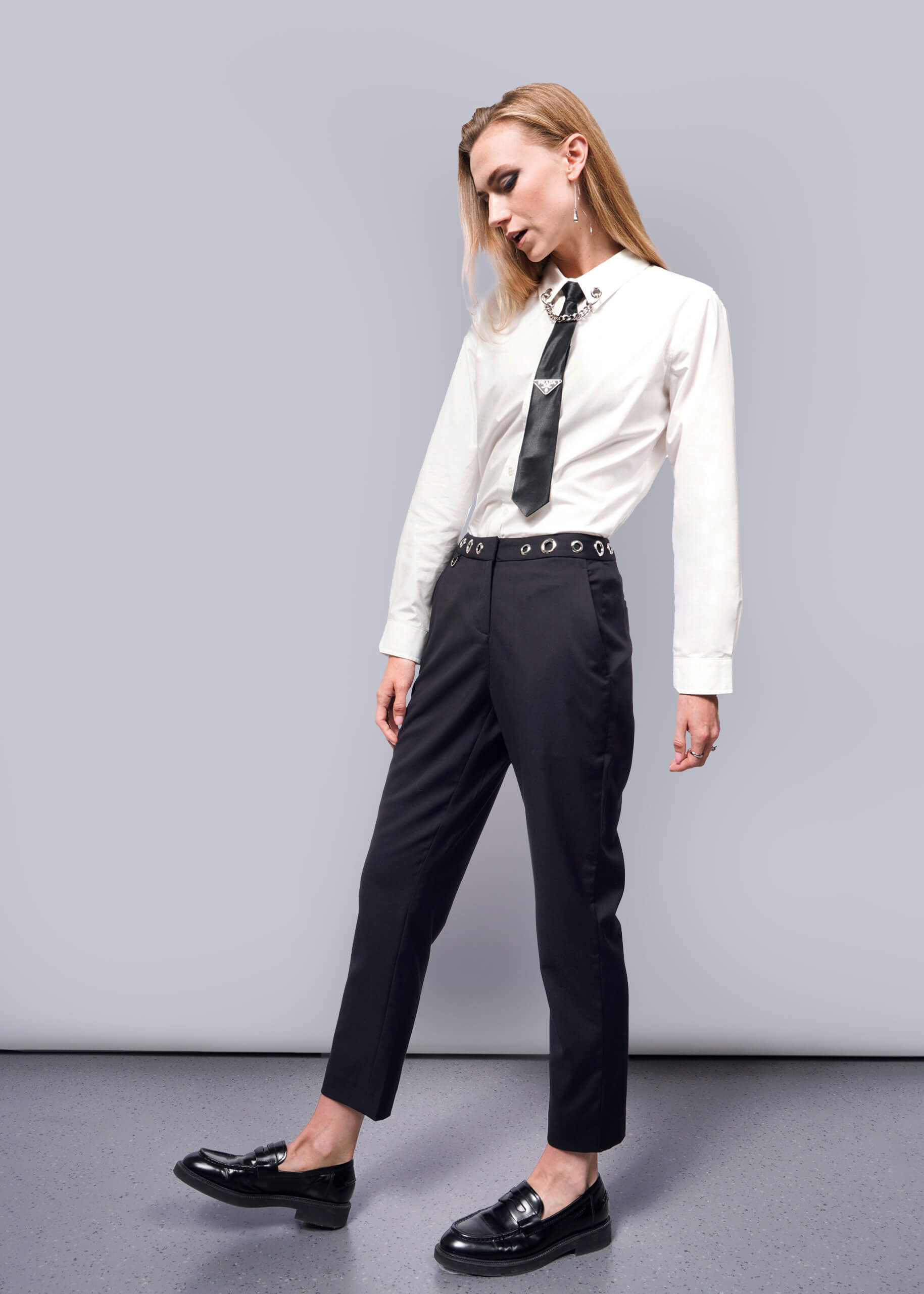 S= A person with long hair channels a punk aesthetic akin to Debbie Harry, standing relaxed against a plain backdrop. They wear a white shirt, black tie, and black loafers from the Wildfang collection, paired with the Debbie Harry X Wildfang Slim Crop Pant. Their head is slightly tilted downwards.