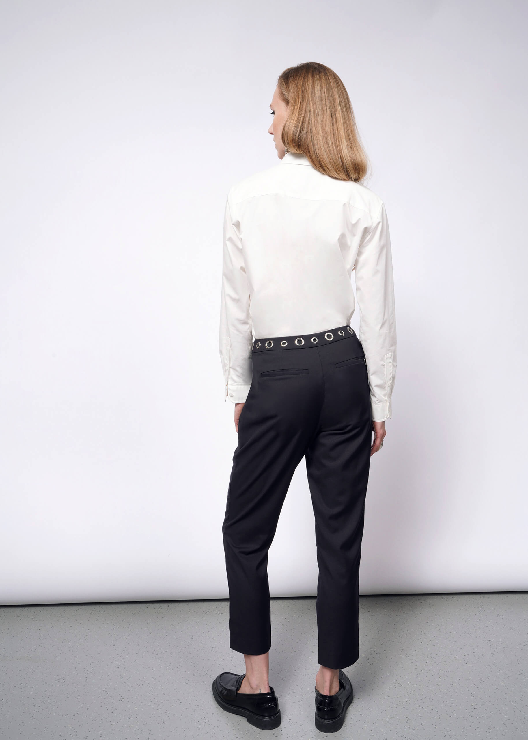 S=A person with long hair, in a white button-up shirt and Debbie Harry X Wildfang Slim Crop Pants featuring a studded belt, stands facing away in a minimalist setting. The gray floor and plain white wall complete the punk-inspired aesthetic of the Wildfang collection.