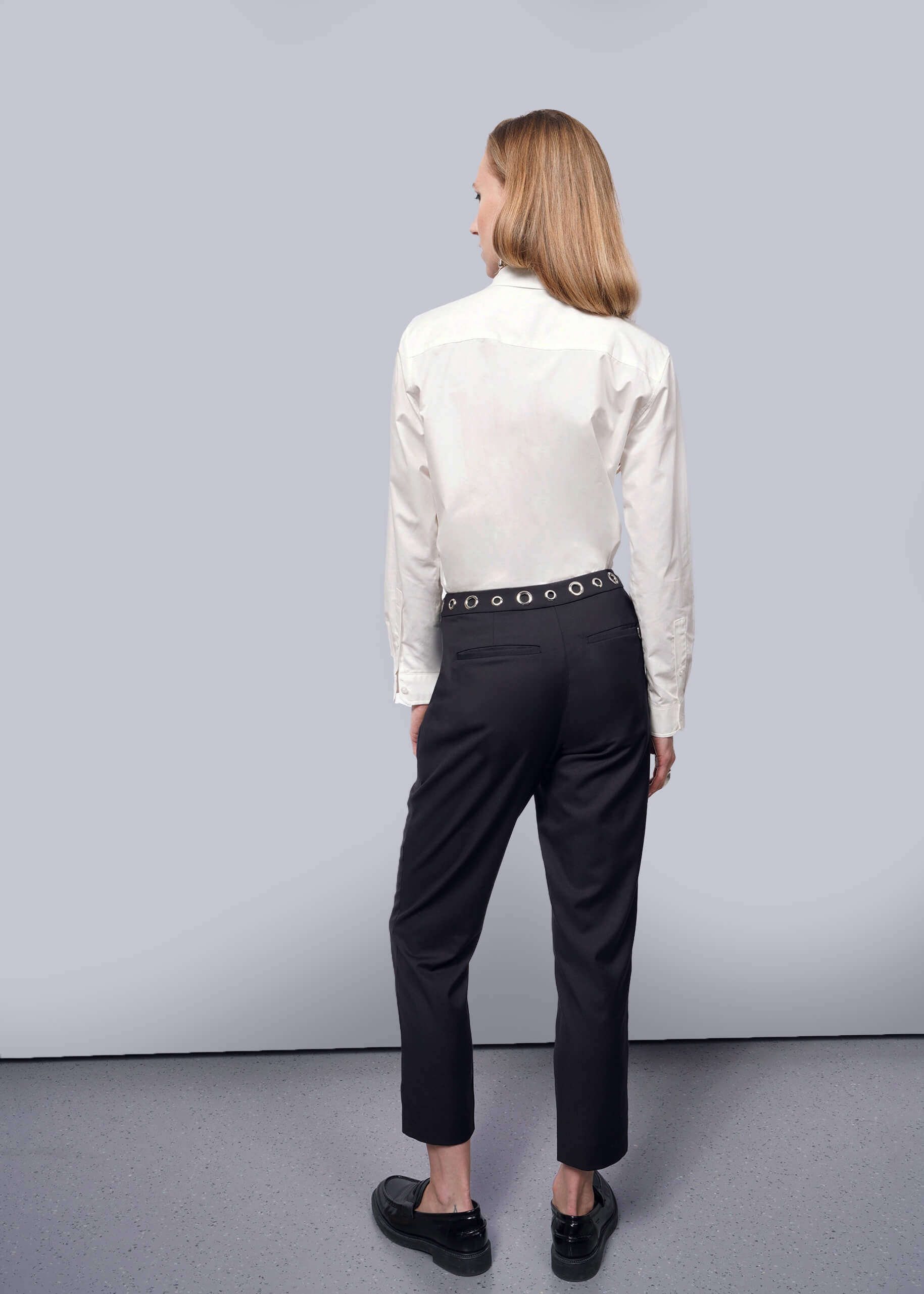 S=A person with long hair, in a white button-up shirt and Debbie Harry X Wildfang Slim Crop Pants featuring a studded belt, stands facing away in a minimalist setting. The gray floor and plain white wall complete the punk-inspired aesthetic of the Wildfang collection.
