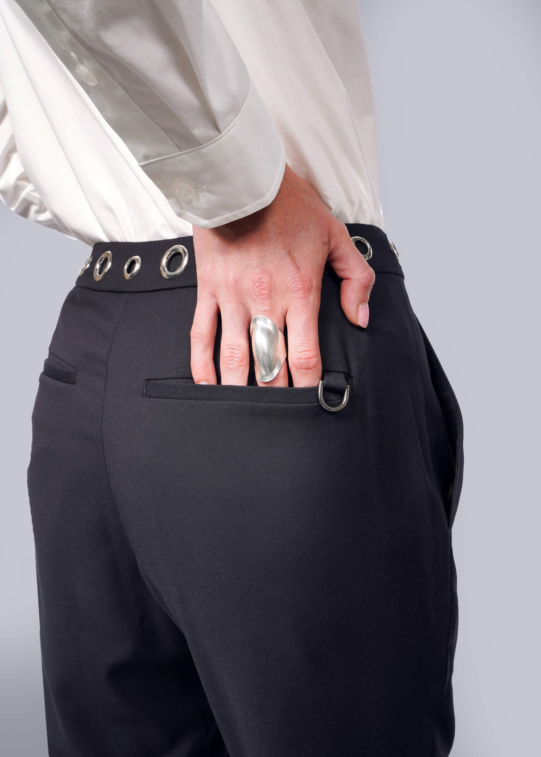 S= A person in a white shirt showcases the back pocket of their Debbie Harry X Wildfang Slim Crop Pant with a grommet belt, delivering a punk vibe. Their hand, featuring a large silver ring in Debbie Harrys iconic style, rests in the pocket, highlighting the ring and pocket detail.
