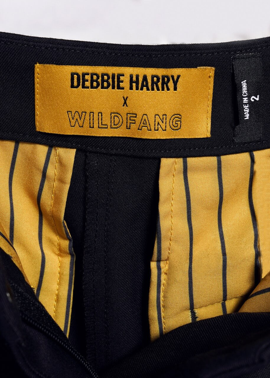1X=Close-up of a clothing label from the Debbie Harry X Wildfang Slim Crop Pant with a punk aesthetic: black text on yellow, yellow and black striped lining, and a bold black size tag with white text on the right.