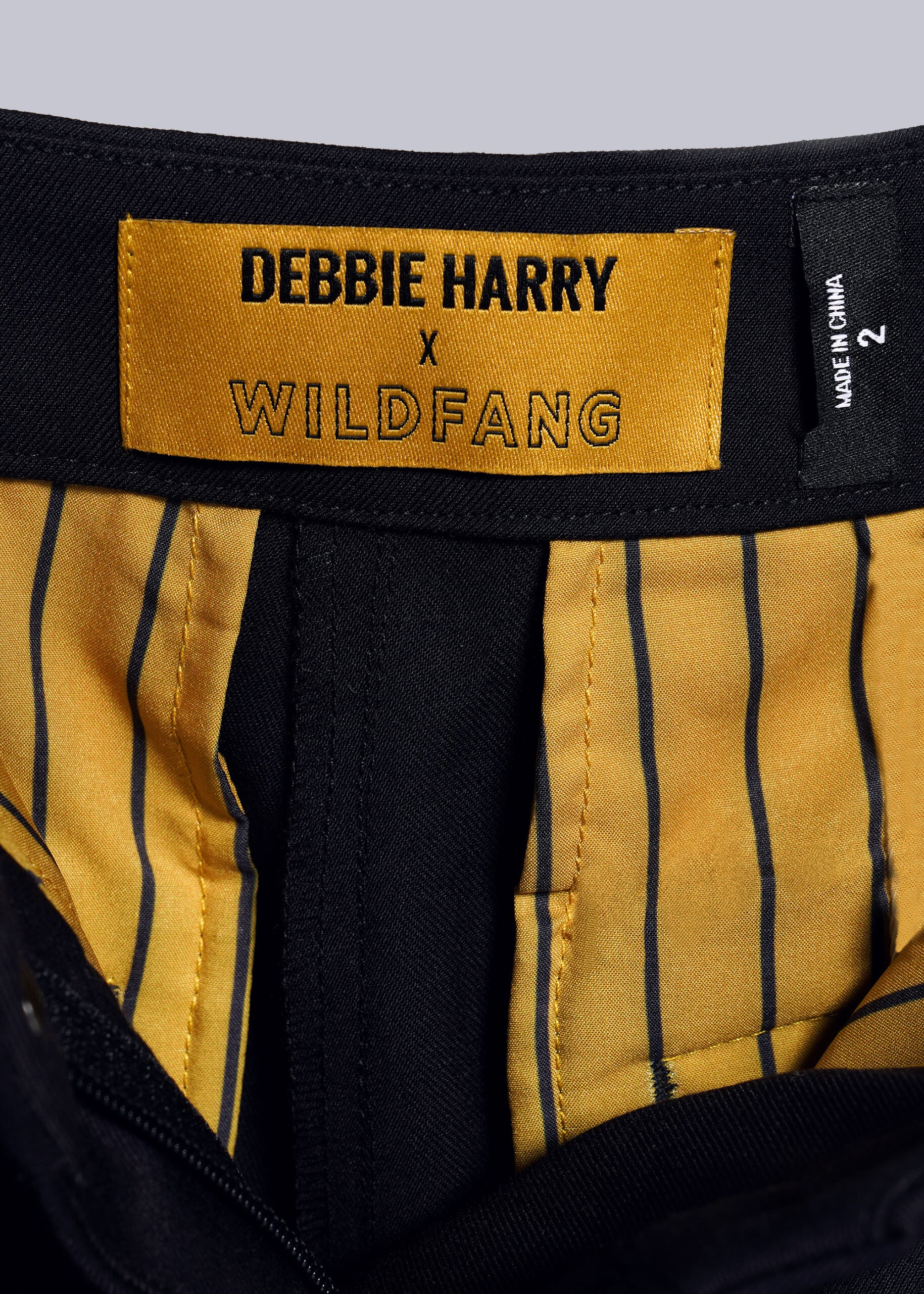 1X=Close-up of a clothing label from the Debbie Harry X Wildfang Slim Crop Pant with a punk aesthetic: black text on yellow, yellow and black striped lining, and a bold black size tag with white text on the right.
