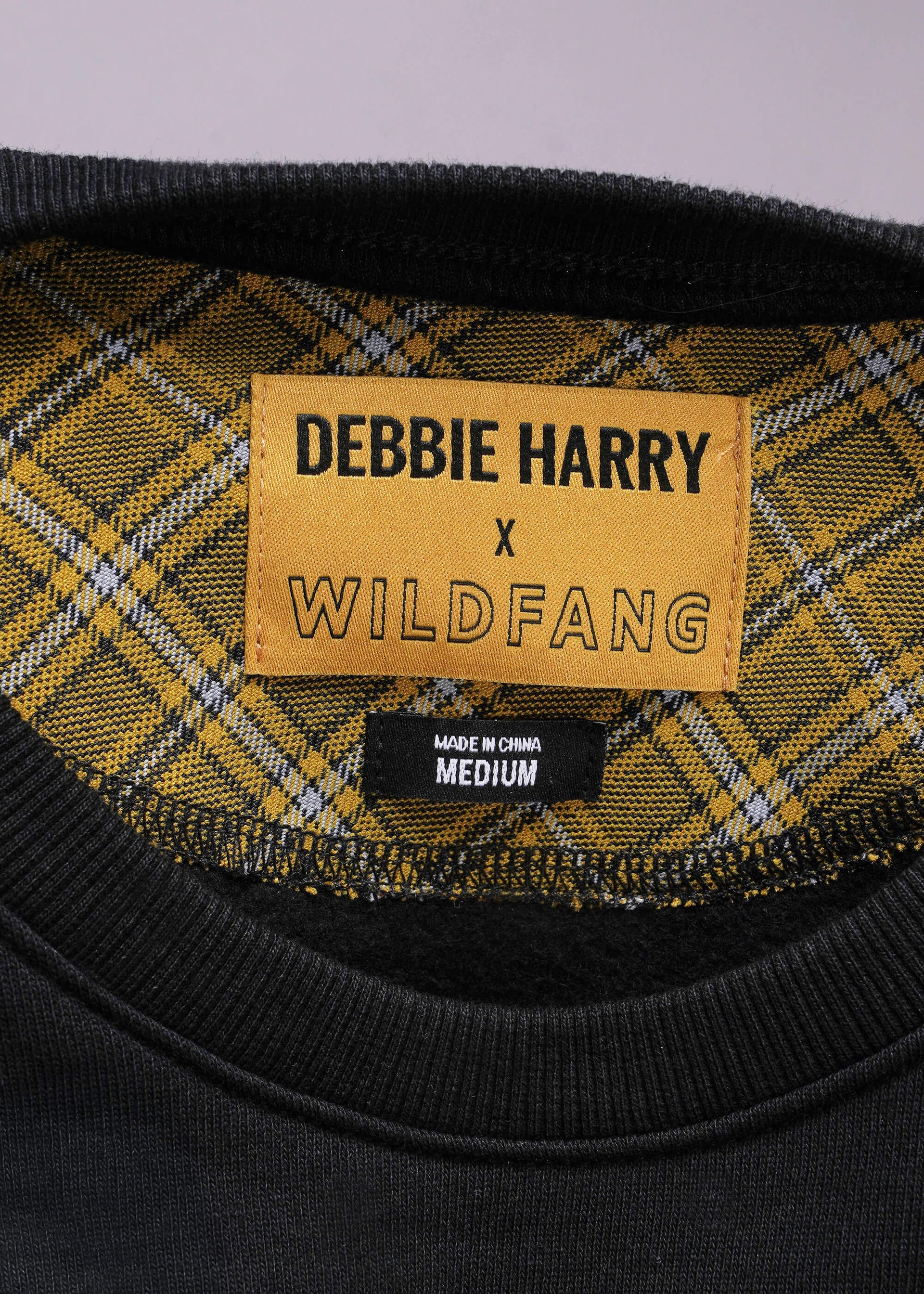 Close-up of a plaid garment with a label reading 'Debbie Harry x Wildfang' on a gray background.