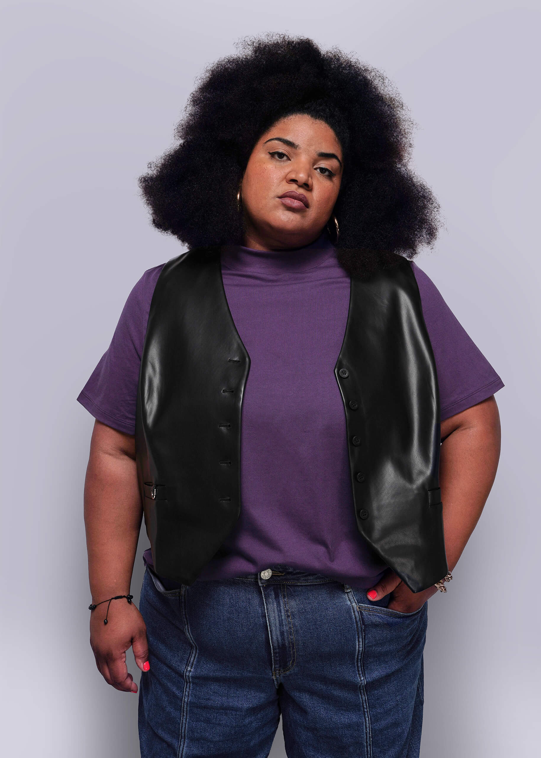 1X=Person wearing a purple shirt, black vegan leather vest, and blue jeans against a light gray background