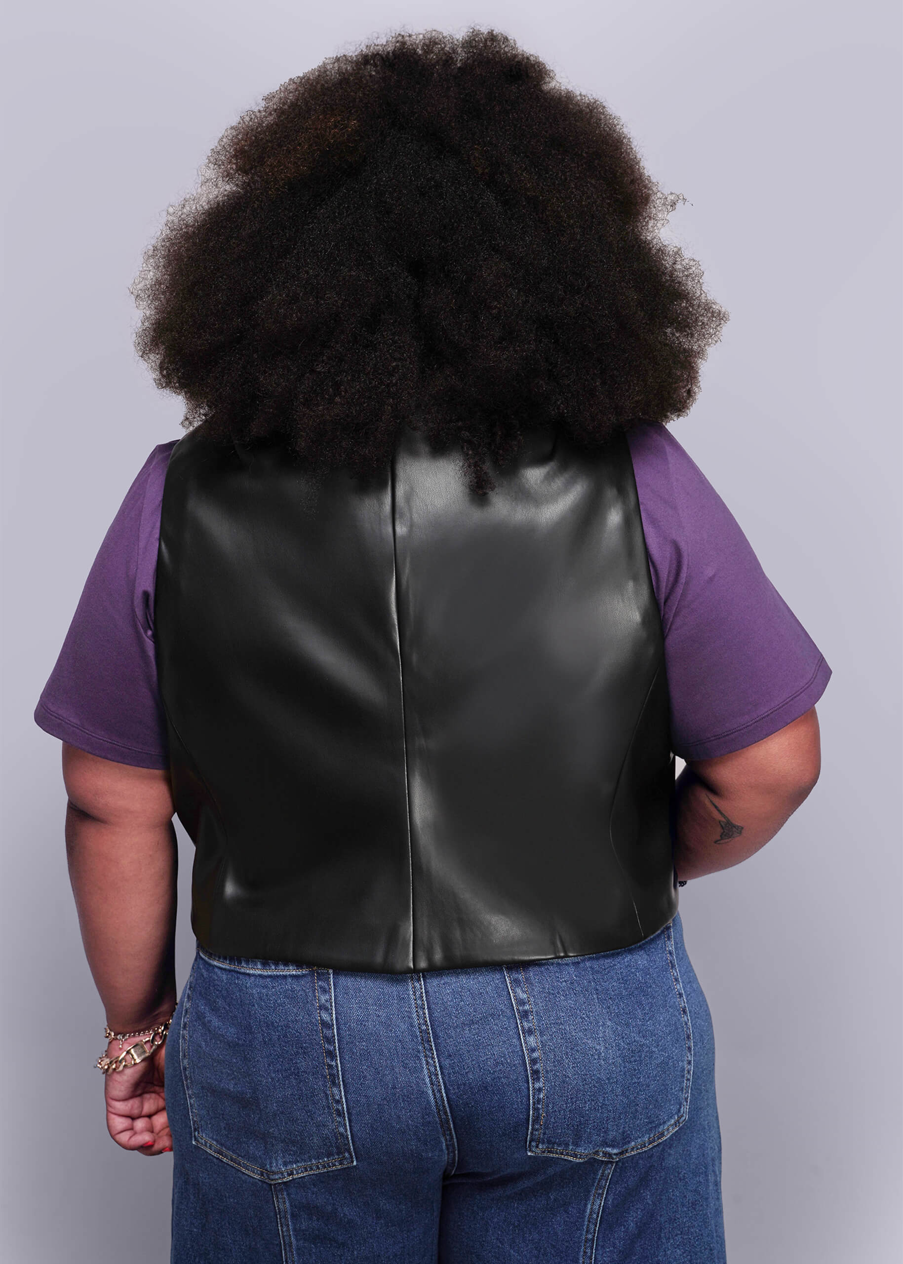 1X=Back view of person wearing a black vegan leather vest over a purple shirt with a plain background