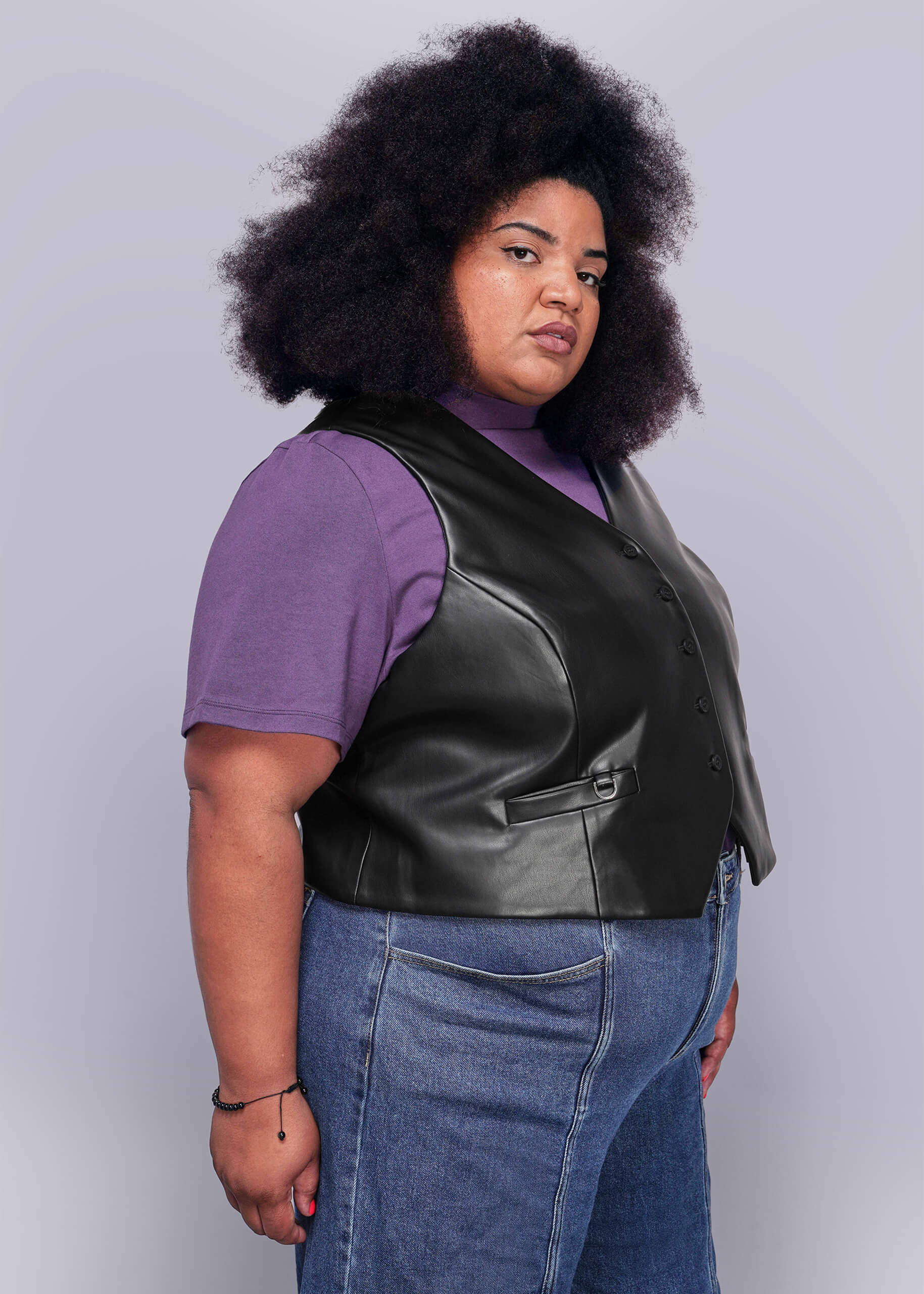 1X=Person wearing a black vegan leather vest over a purple shirt with blue jeans on a light gray background