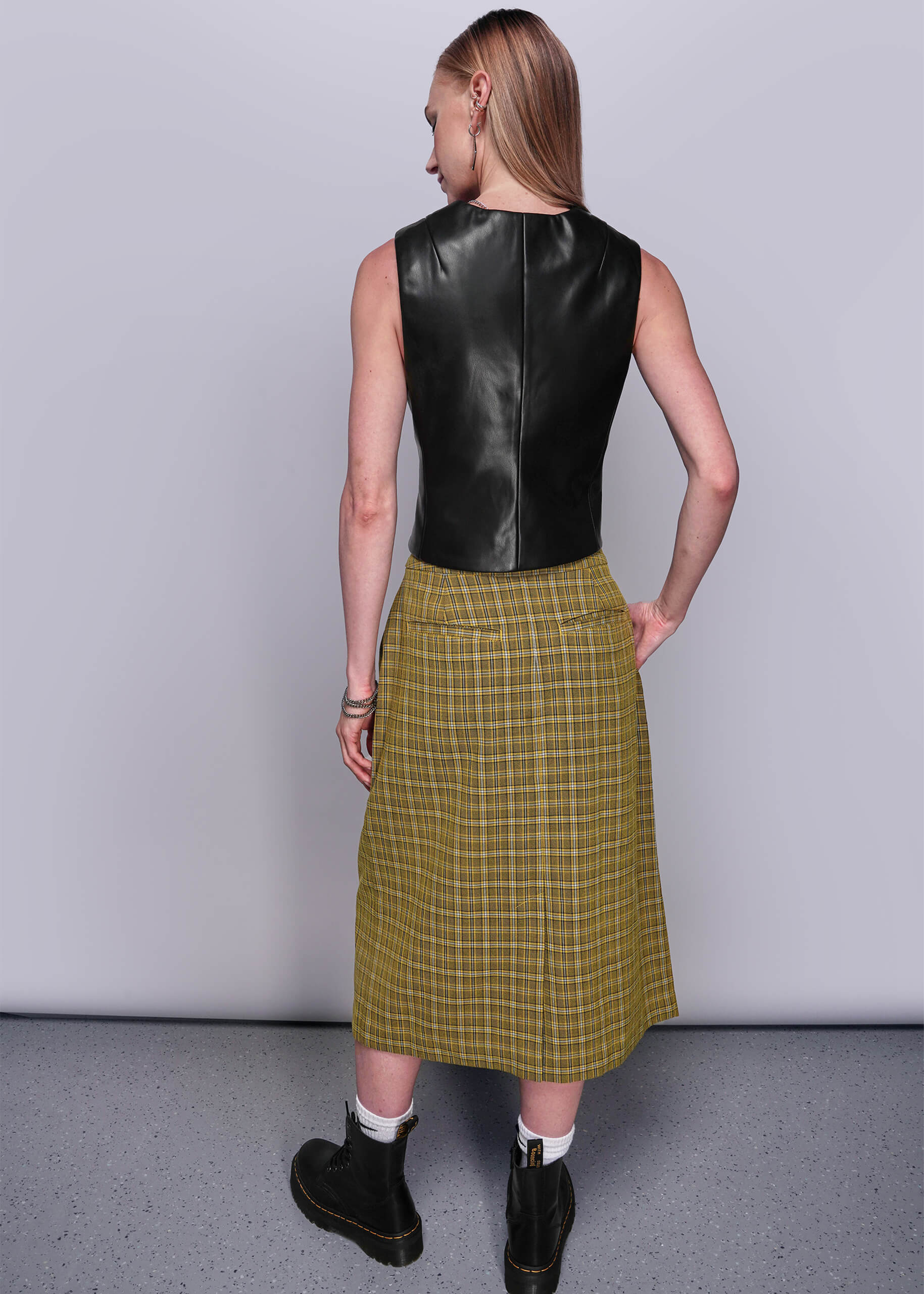 S=Person wearing a black vegan leather vest and yellow plaid skirt against a plain background