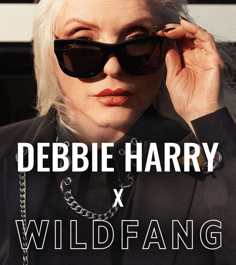 Close up of Debbie Harry looking at camera wearing sunglasses and pieces from the Debbie Harry x Wildfang collection. Text overlay says DEBBIE HARRY X WILDFANG.