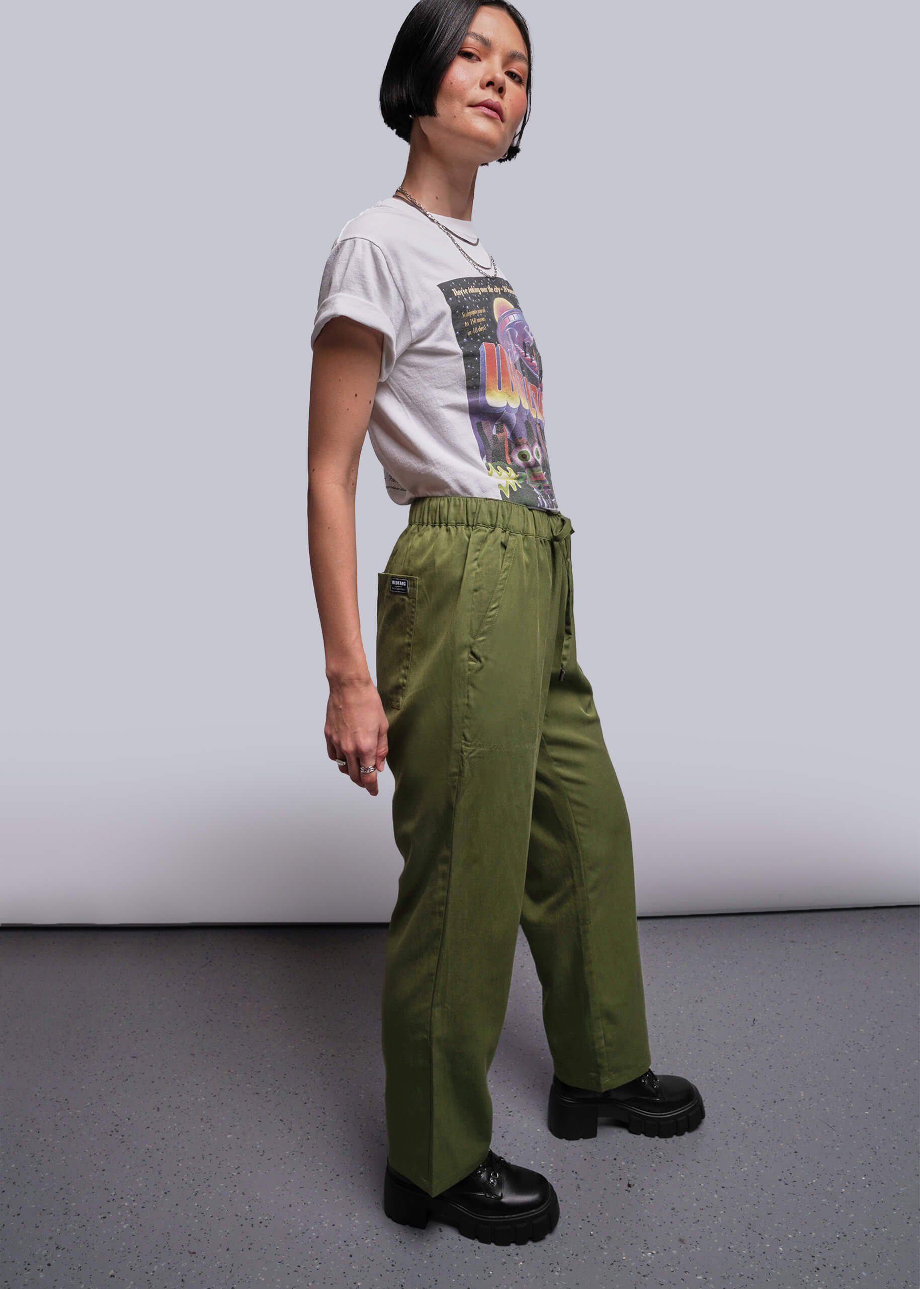 S=Person wearing a white t-shirt with colorful graphics and green pants against a plain background