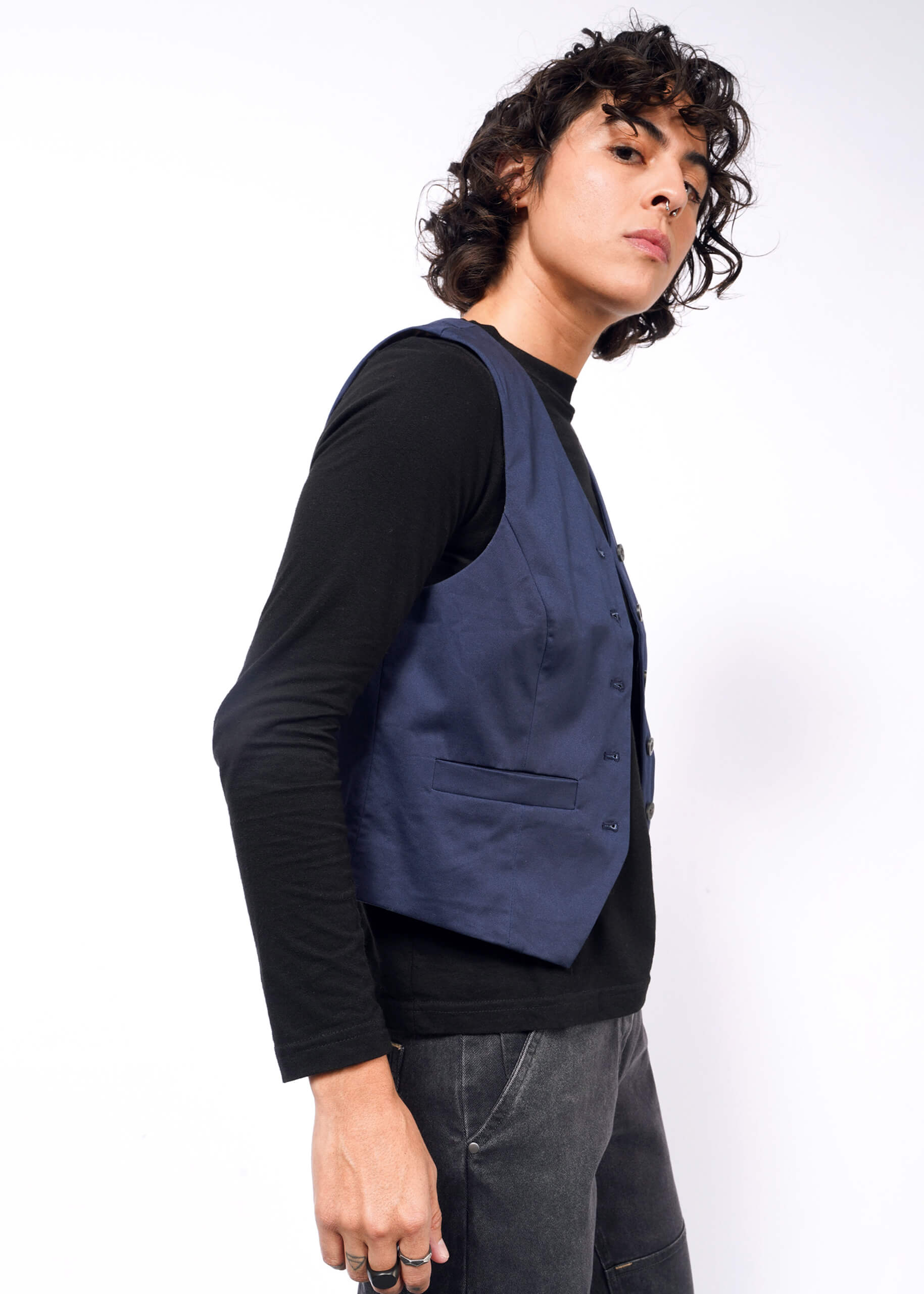 S=A person with curly hair wears The Essential Vest in blue over a black long-sleeve shirt and dark jeans, standing against a white background.