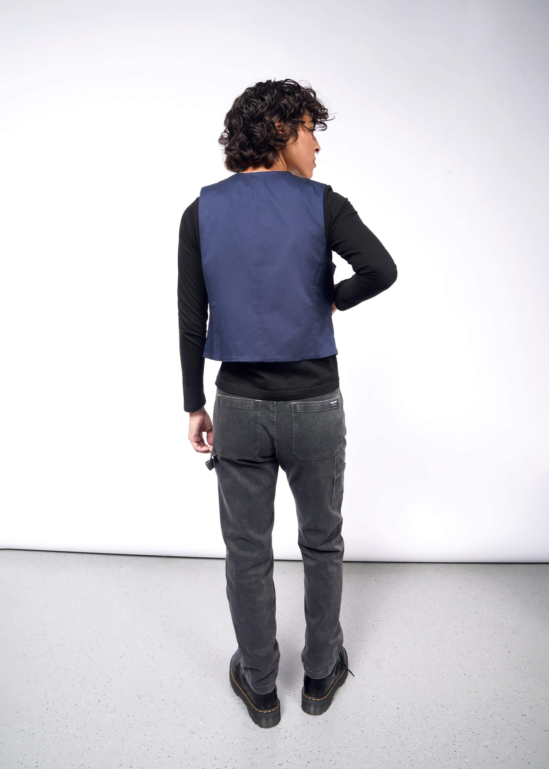 S=A man with curly hair is wearing a black long-sleeve shirt, The Essential Vest tailored fit in stretch cotton, and gray jeans, standing on a light gray floor facing a white wall.