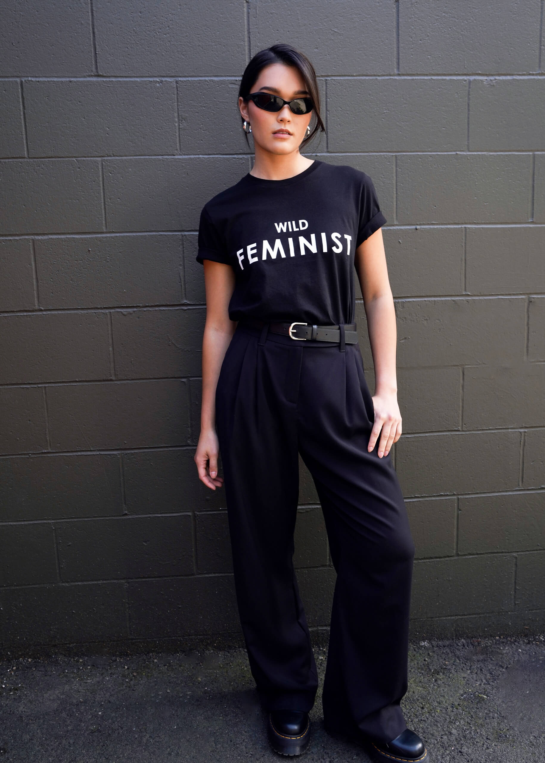S=Against a dark brick wall, a person exudes confidence, wearing trendy sunglasses and The Wild Feminist® Tee—a black cotton unisex fit t-shirt with WILD FEMINIST printed on it—paired with black high-waisted pants and matching shoes. With one hand in their pocket, they embody style.