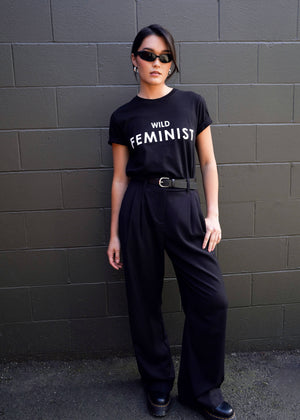 S=Against a dark brick wall, a person exudes confidence, wearing trendy sunglasses and The Wild Feminist® Tee—a black cotton unisex fit t-shirt with WILD FEMINIST printed on it—paired with black high-waisted pants and matching shoes. With one hand in their pocket, they embody style.