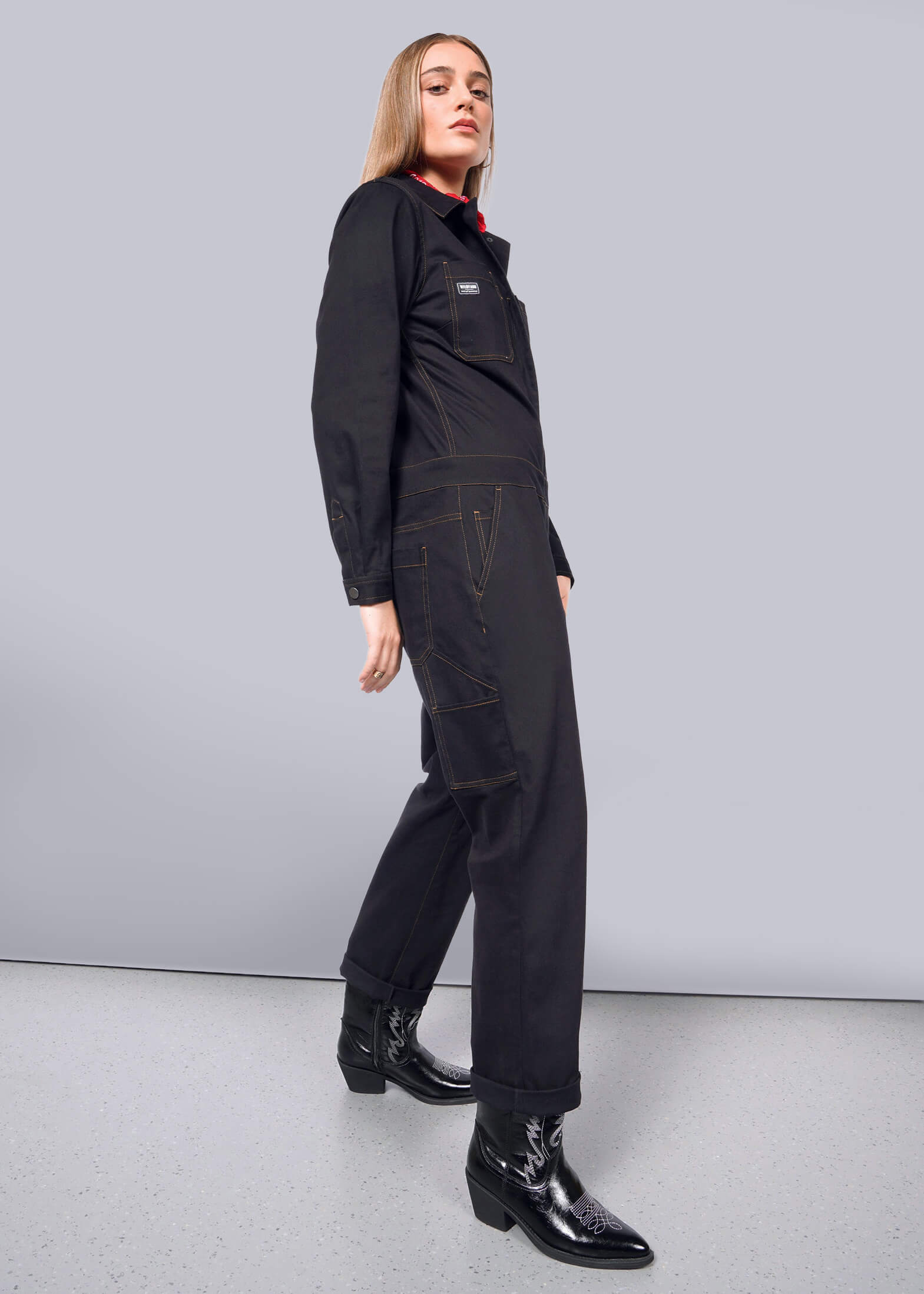 S=Against a white wall, the person exudes confidence in The Essential Long Sleeve Coverall and shiny boots. Long hair frames their face as they gaze into the camera, embodying a bold style.