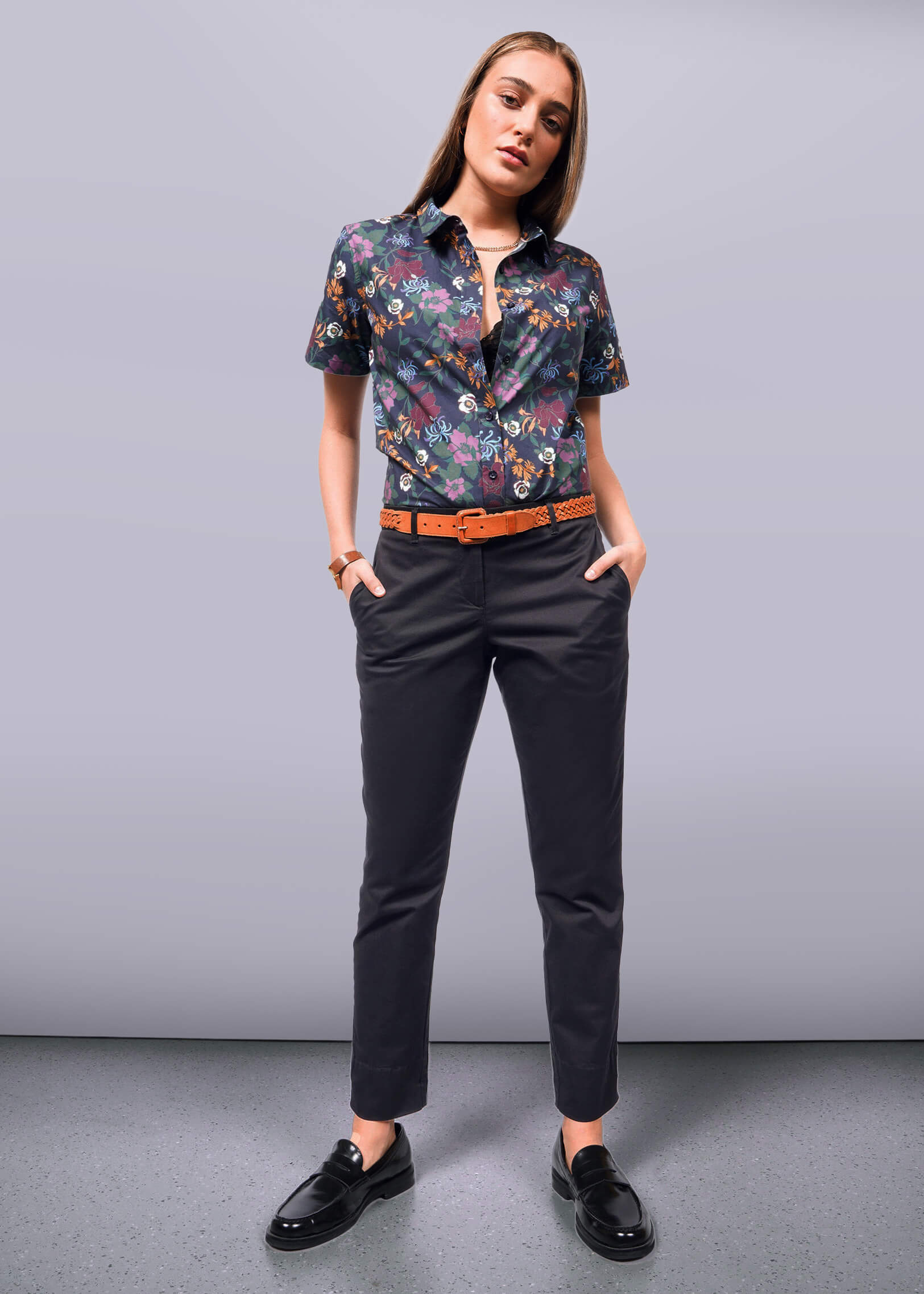 S=The woman stands confidently in a floral short-sleeve shirt and The Essential Trouser in relaxed mid-rise black. Black loafers and a brown belt with a bracelet complete the look. Her hands rest in her pockets against a plain light gray wall and floor, highlighting her poised stance.
