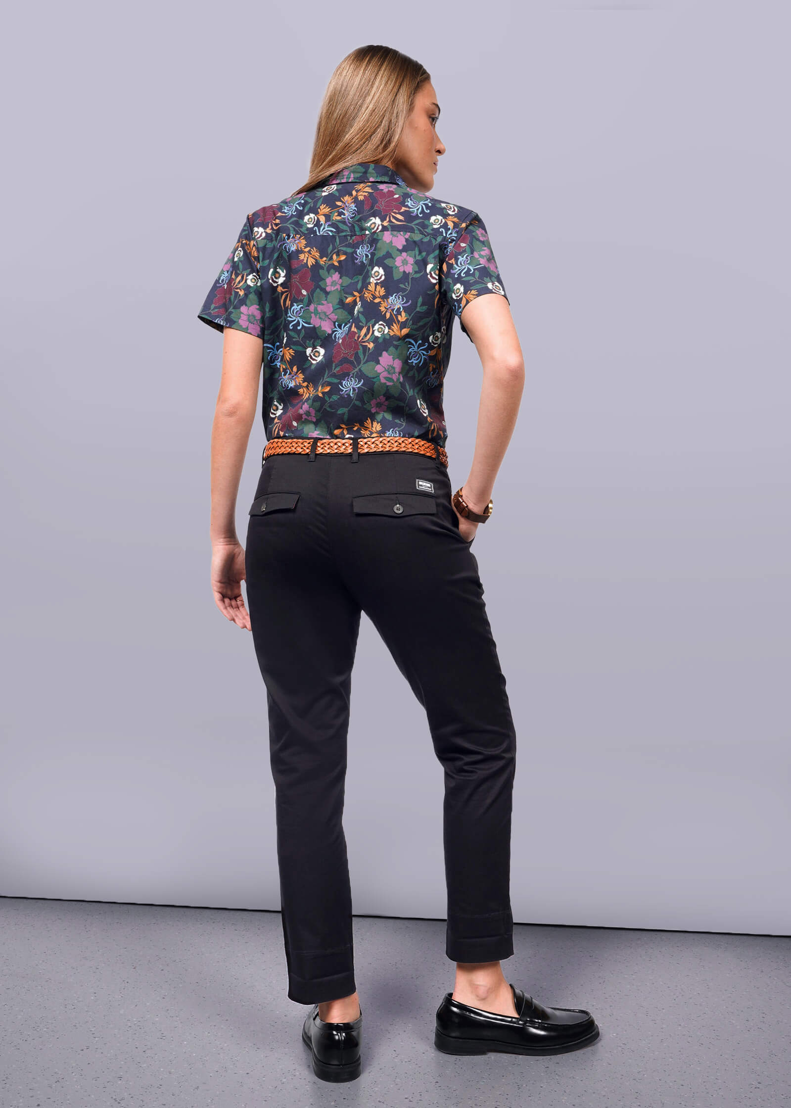 S=A person with long hair stands facing away, showcasing a floral short-sleeve shirt and The Essential Trouser in black with matching shoes. They stand on a light gray floor against a plain white background.