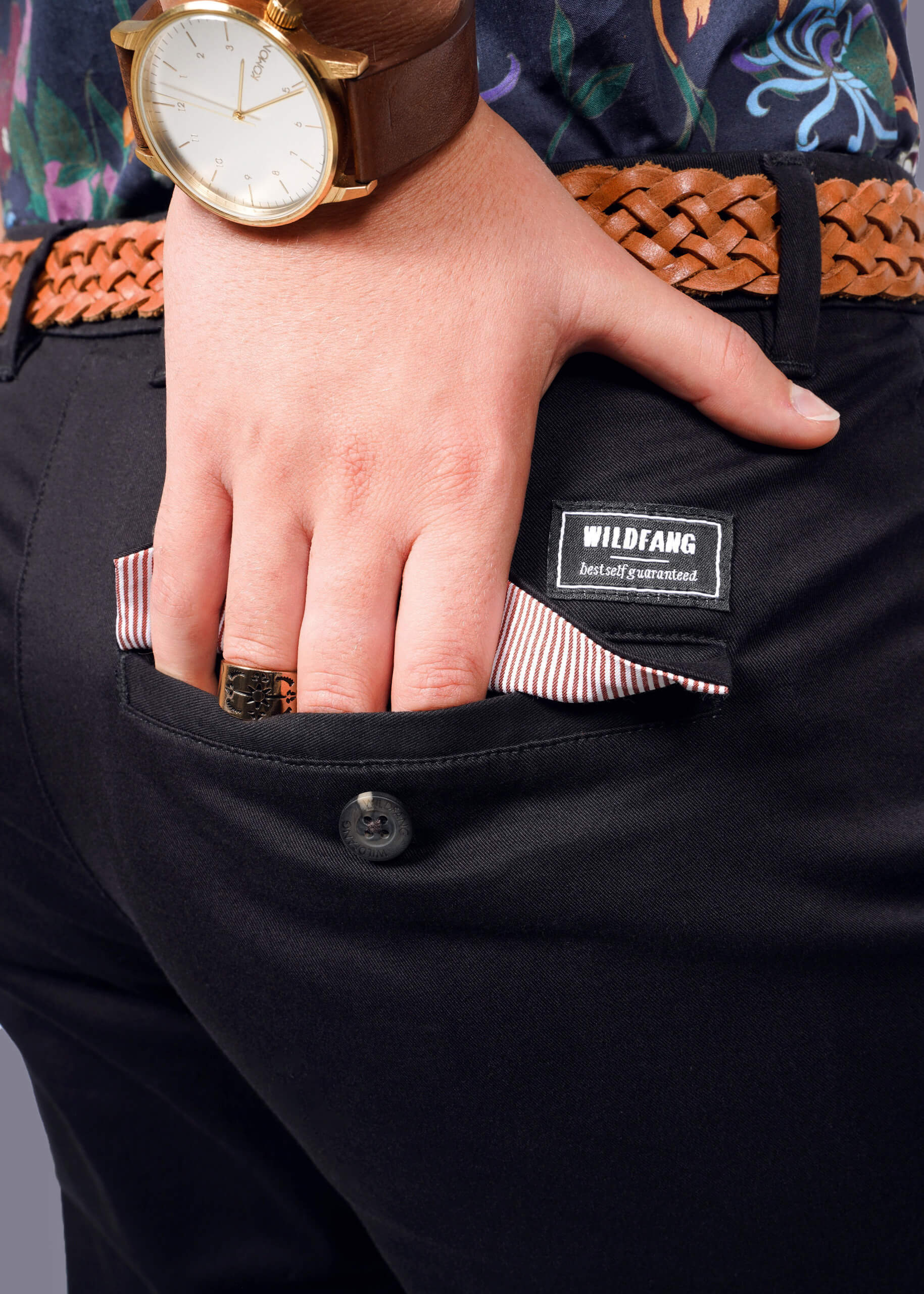 S=A close-up shows someone in The Essential Trouser, featuring a relaxed mid-rise fit and dark finish, accented by a woven brown belt. A WILDFANG tag is visible as their hand rests in a pocket lined with red and white stripes. They wear a watch with a brown leather strap and a ring for a timeless look.