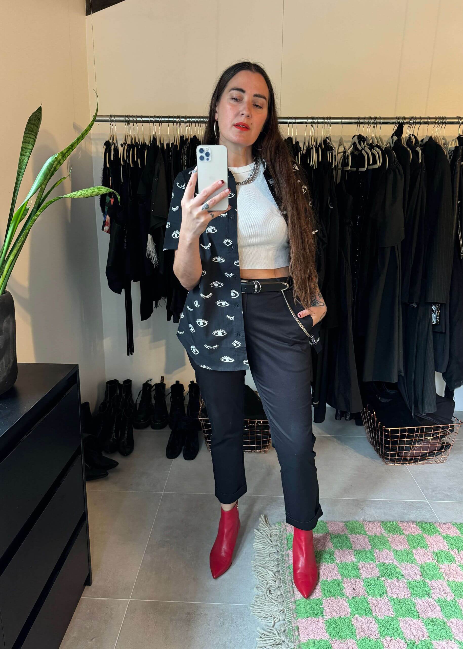 A woman takes a mirror selfie in a room with clothing racks. She wears The Essential Trouser in black and white paired with red boots, showcasing a relaxed mid-rise fit. Shes holding a phone, with a plant and rug completing the scene.