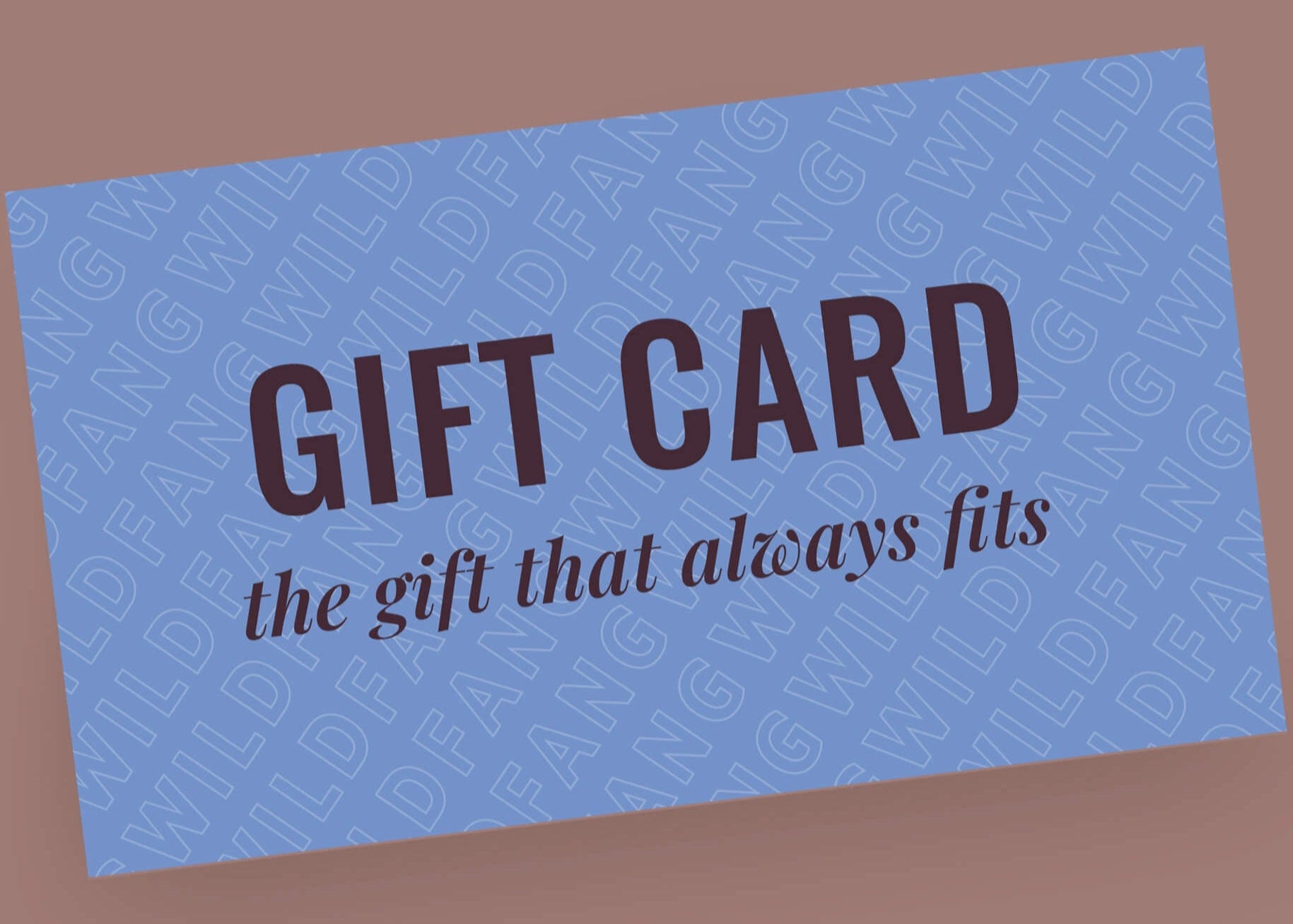 A blue Gift Card, labeled the gift that always fits, features bold black GIFT CARD text and a repeating WILDFANG background. Resting on a mauve surface, these climate-neutral cards make an ideal gift for any occasion.