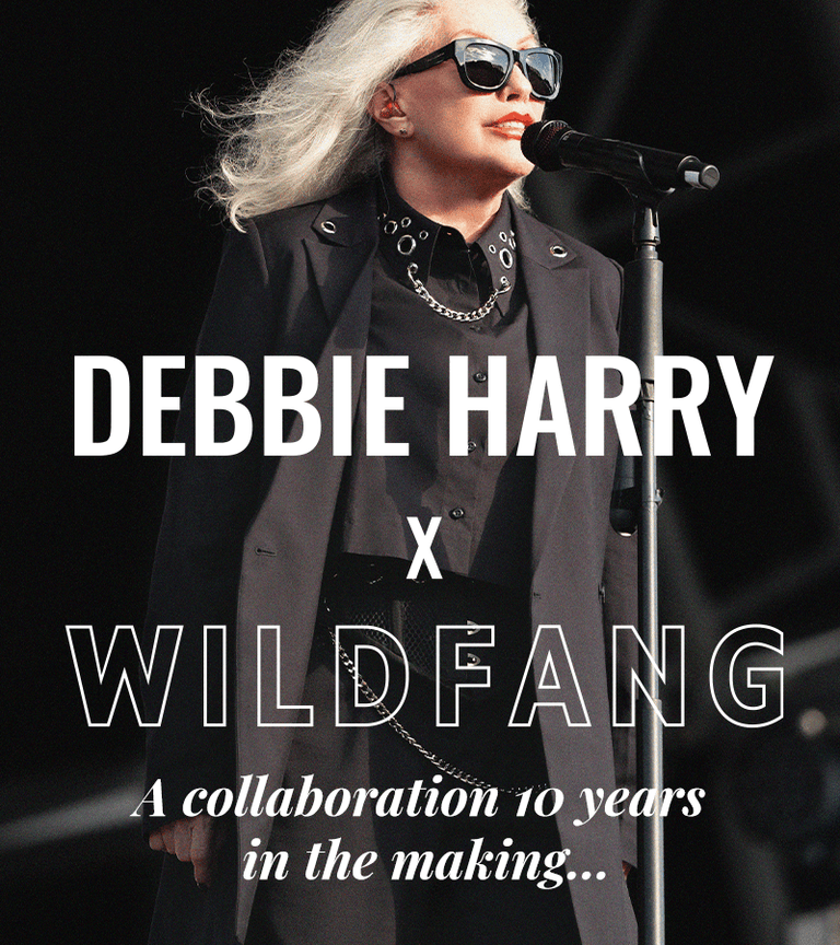Close up of Debbie Harry with microphone stand on stage at her concert. She wears sunglasses and a suit and button up from the Debbie Harry x Wildfang collection. Text overlay centered and towards bottom of image reads "DEBBIE HARRY X WILDFANG, a collaboration 10 years in the making..."