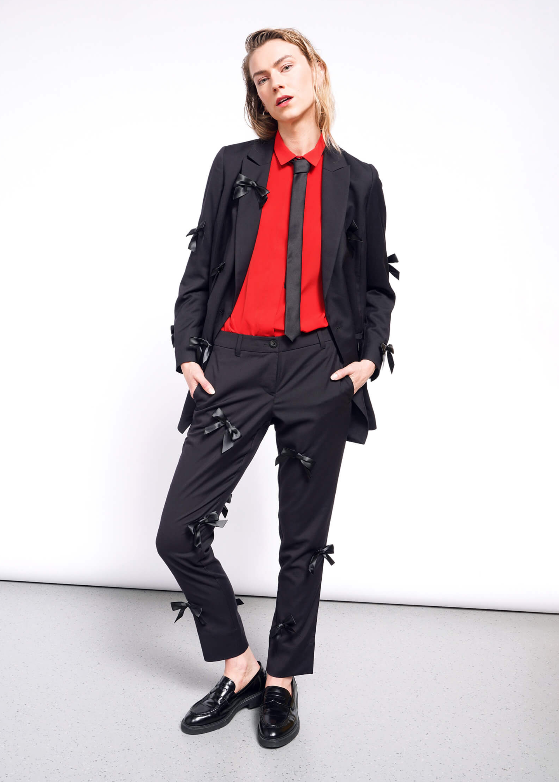 S=A person poses against a white background wearing a black suit with The Empower Bows Trouser, bow-embellished pants, a red shirt, black tie, and loafers. One hand rests in their pocket, exuding an ultra-luxe feel.