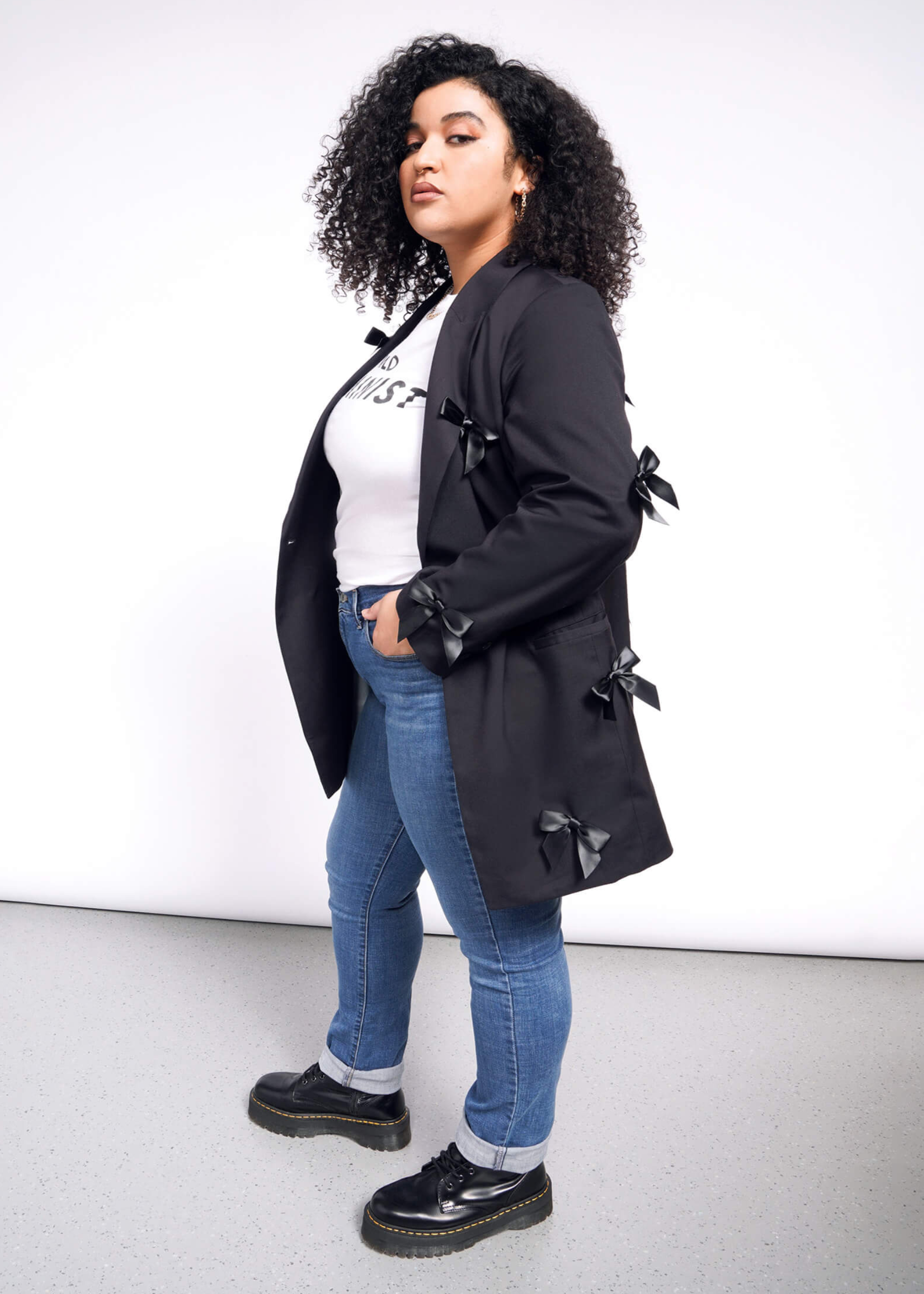 1X=A person with curly hair stands confidently in a white t-shirt, blue jeans, and black shoes, wearing The Empower Bows Double Breasted Blazer adorned with decorative side bows. The background is a plain mix of white and gray.