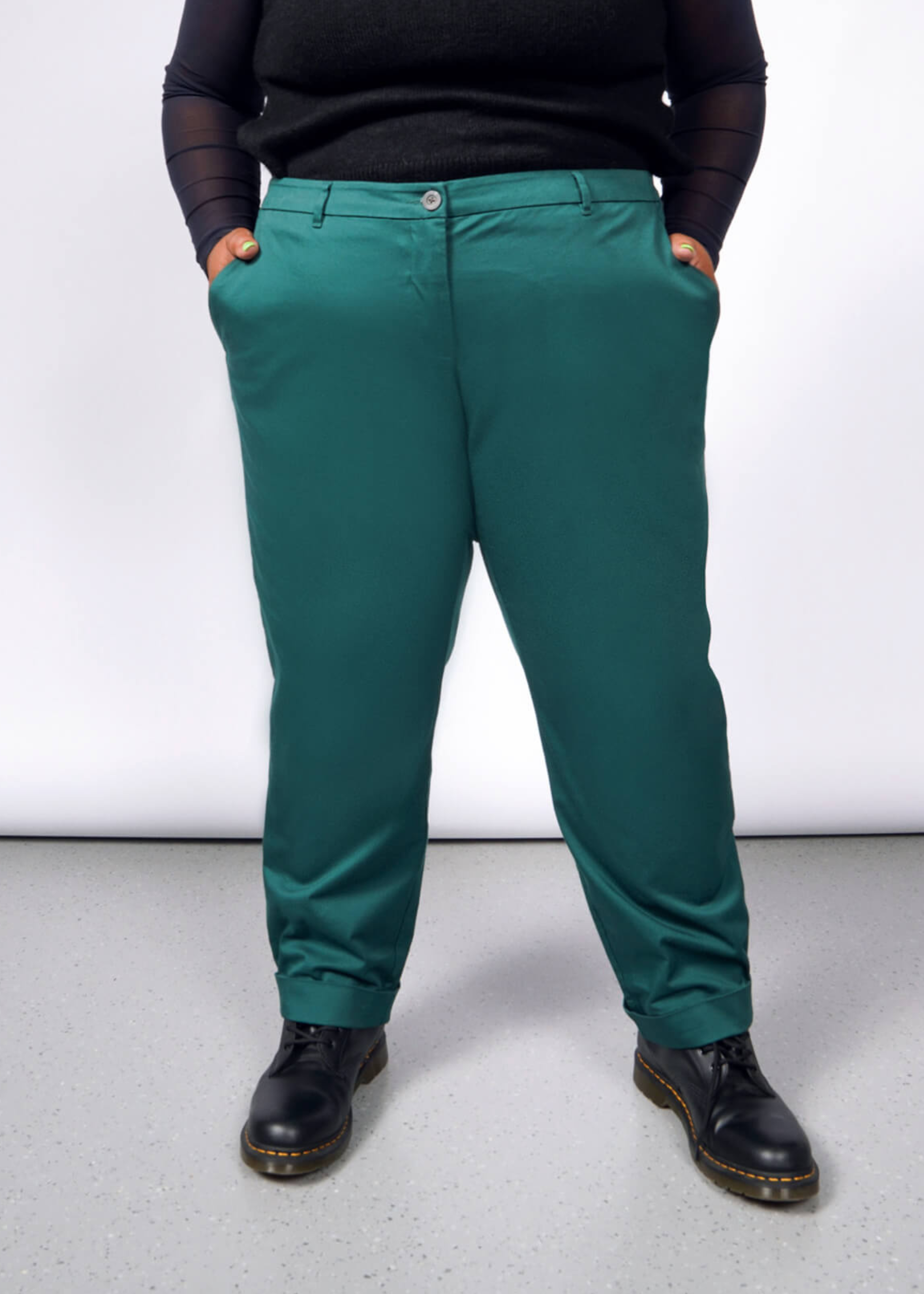 1X= A person models The Essential Trouser in classic teal with black shoes, standing confidently with hands in pockets against a light gray backdrop, highlighting the timeless wardrobe staple.