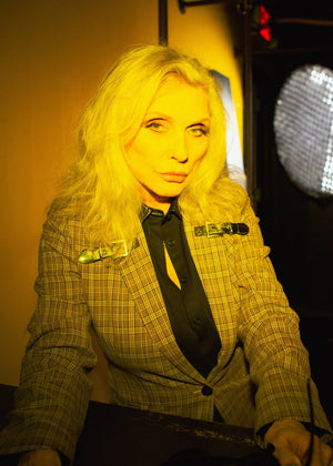 Person with blonde hair wearing a checkered suit in a dimly lit room.