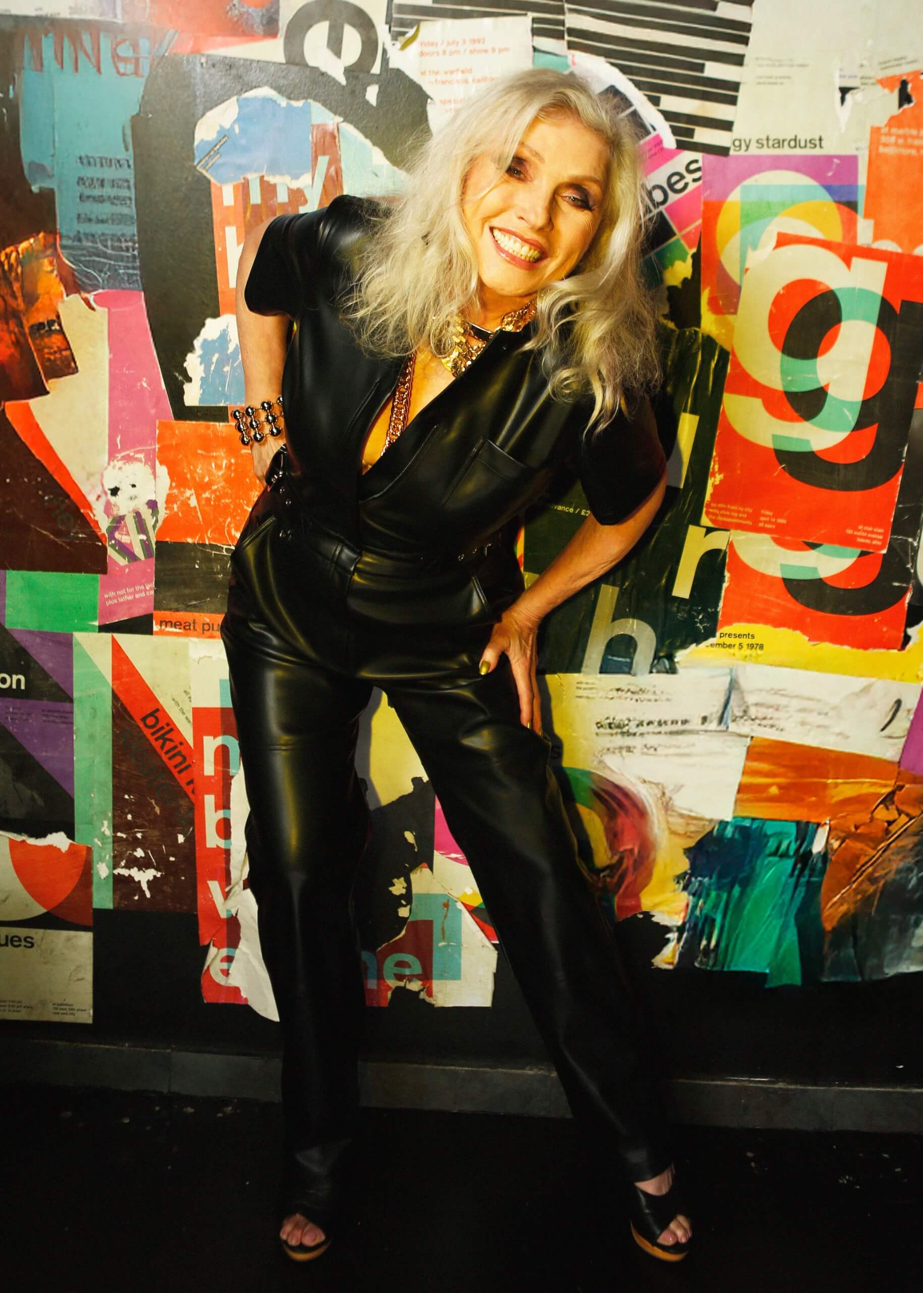 Debbie Harry wears the Debbie Harry x Wildfang Vegan Leather coverall standing in front of a colorful collage wall.