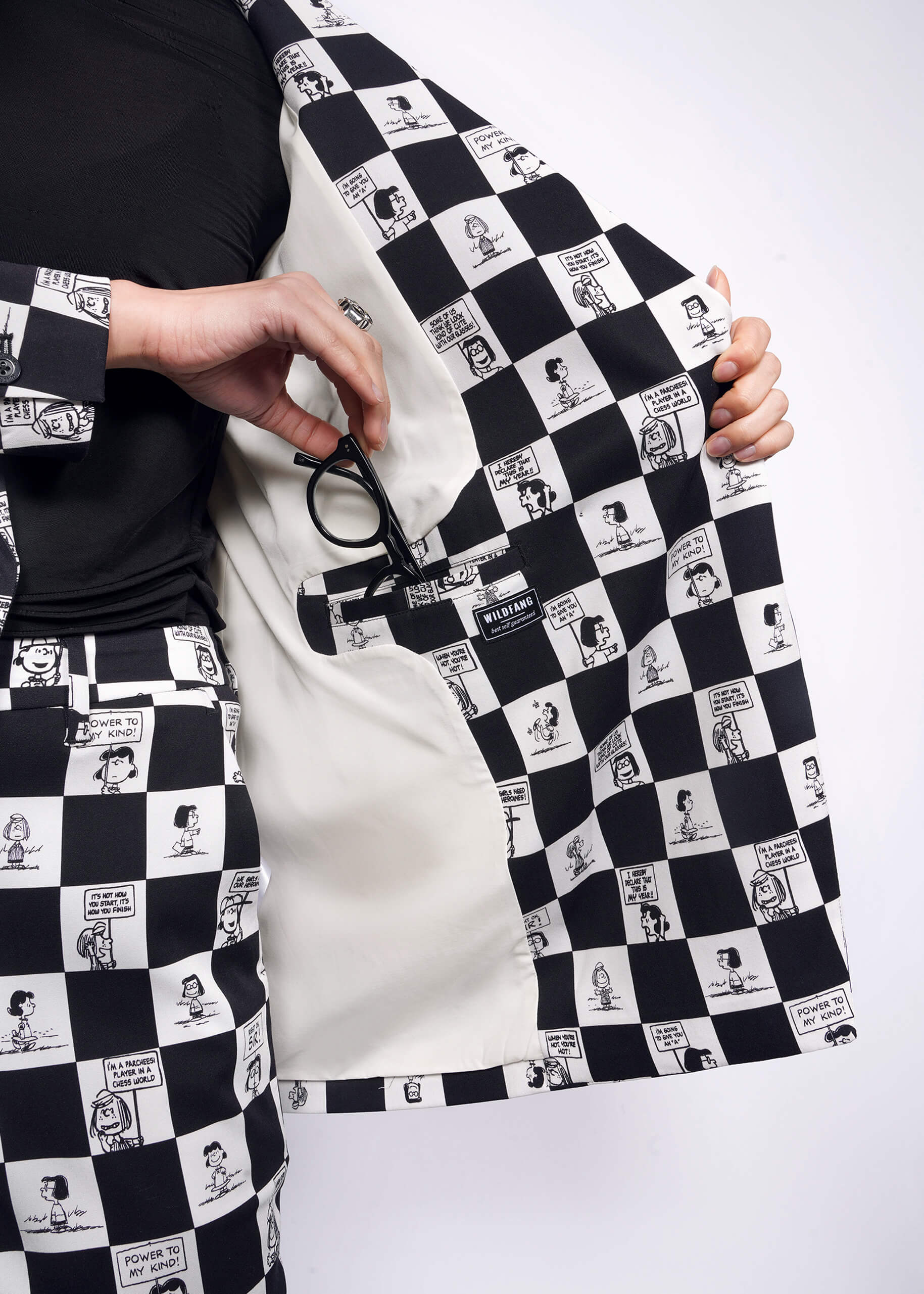S=Close up view of model putting sunglasses in interior blazer pocket of The Peanuts x Wildfang Double Breasted Blazer in a custom black and white checkered print featuring Peanuts characters.